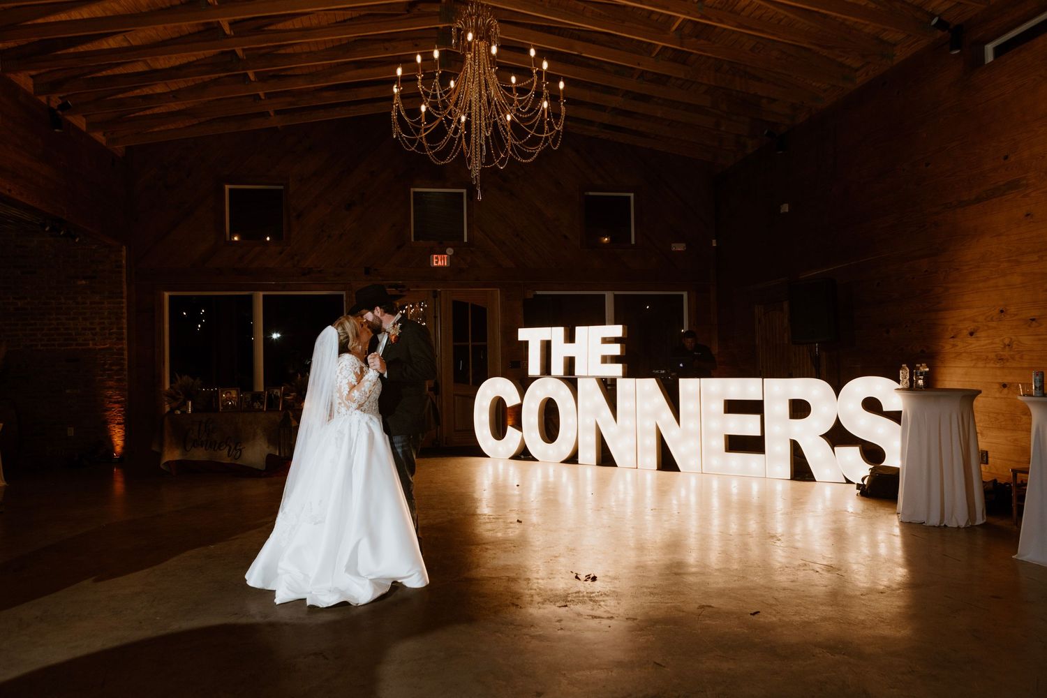 Bride and groom share first dance before large marquee letters spelling THE CONNERS in a rustic wedding venue.