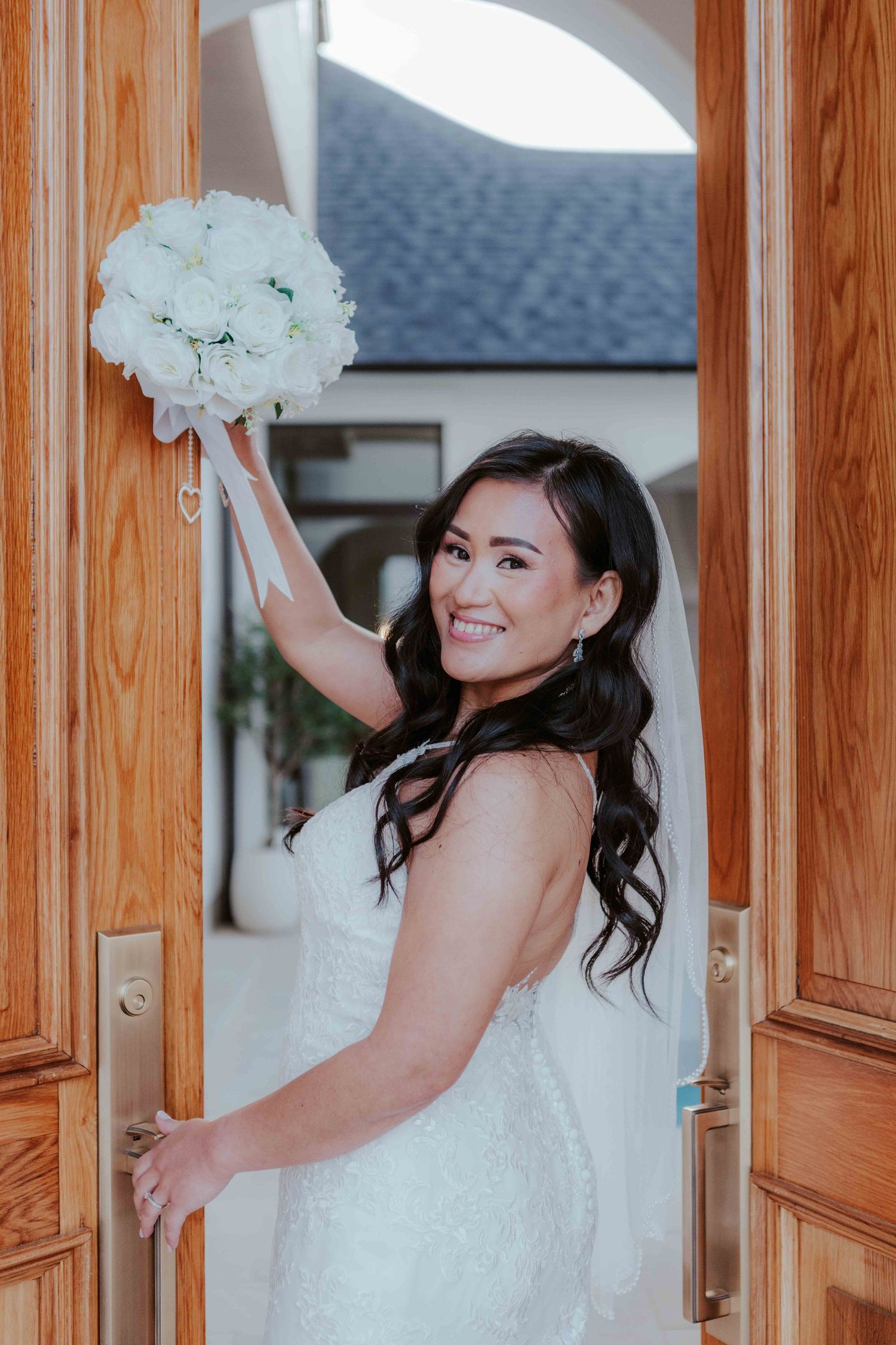 A bride in a white lace wedding dress holds up a white floral bouquet while standing in a wooden doorway.