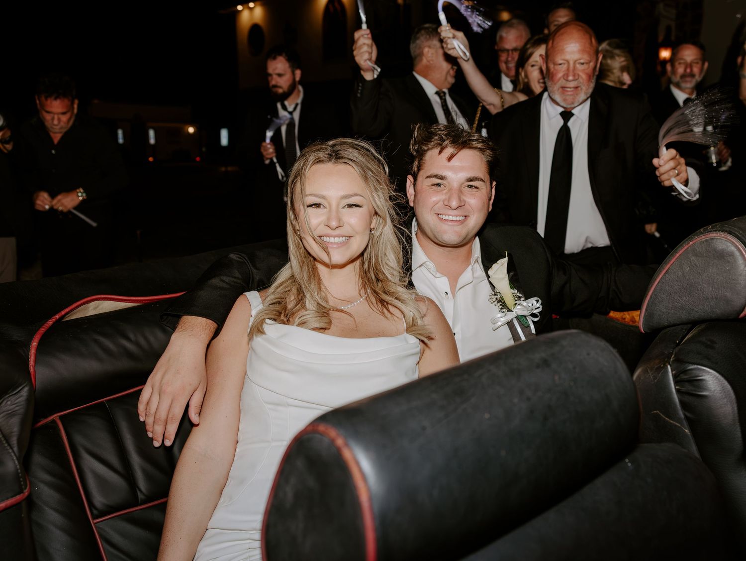 Smiling couple in white dress and dark suit with boutonniere sit on black leather theater seats as guests clap behind them.