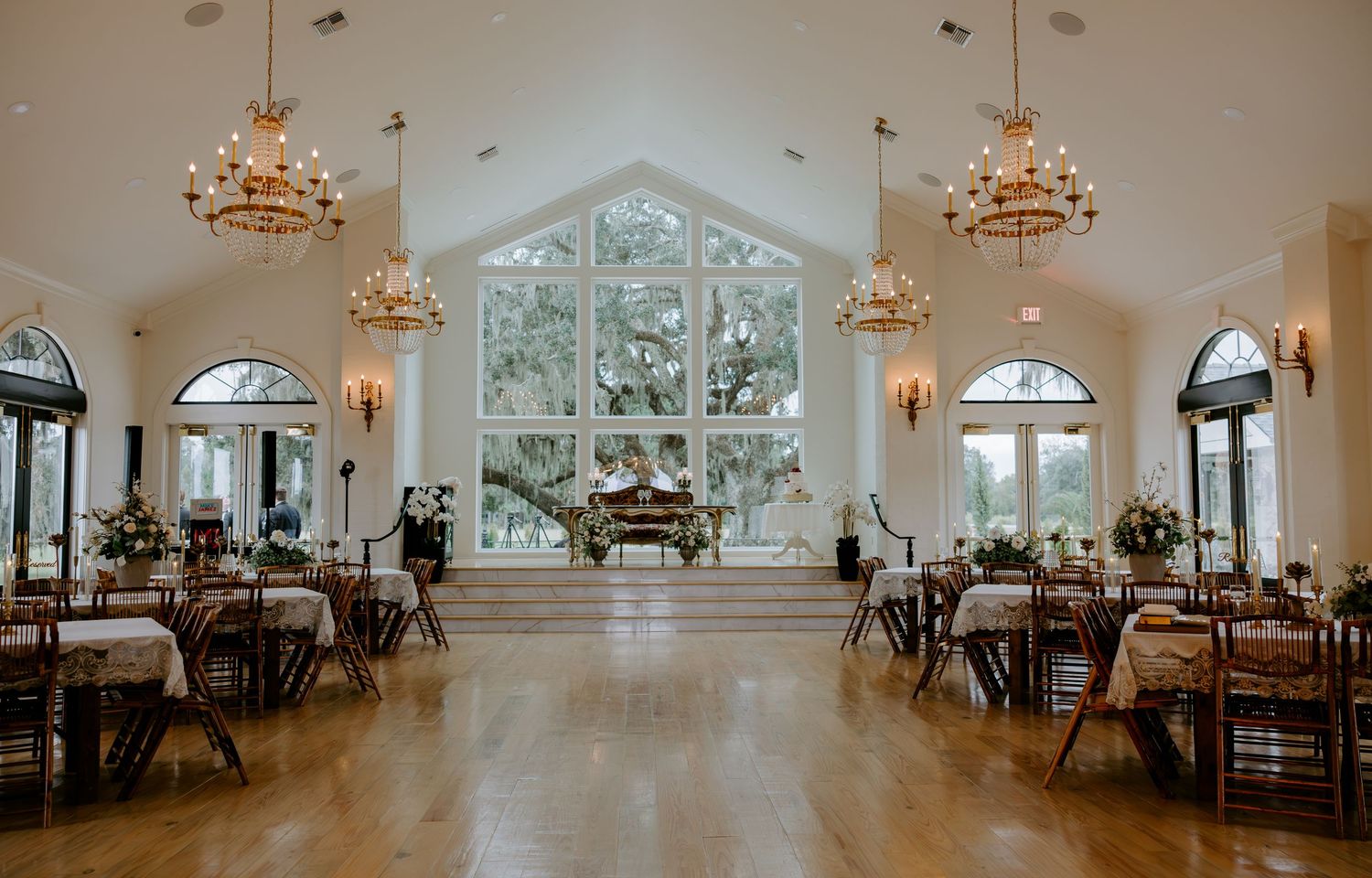 Elegant wedding venue interior with chandeliers, hardwood floors, banquet tables, and tall arched windows overlooking trees.