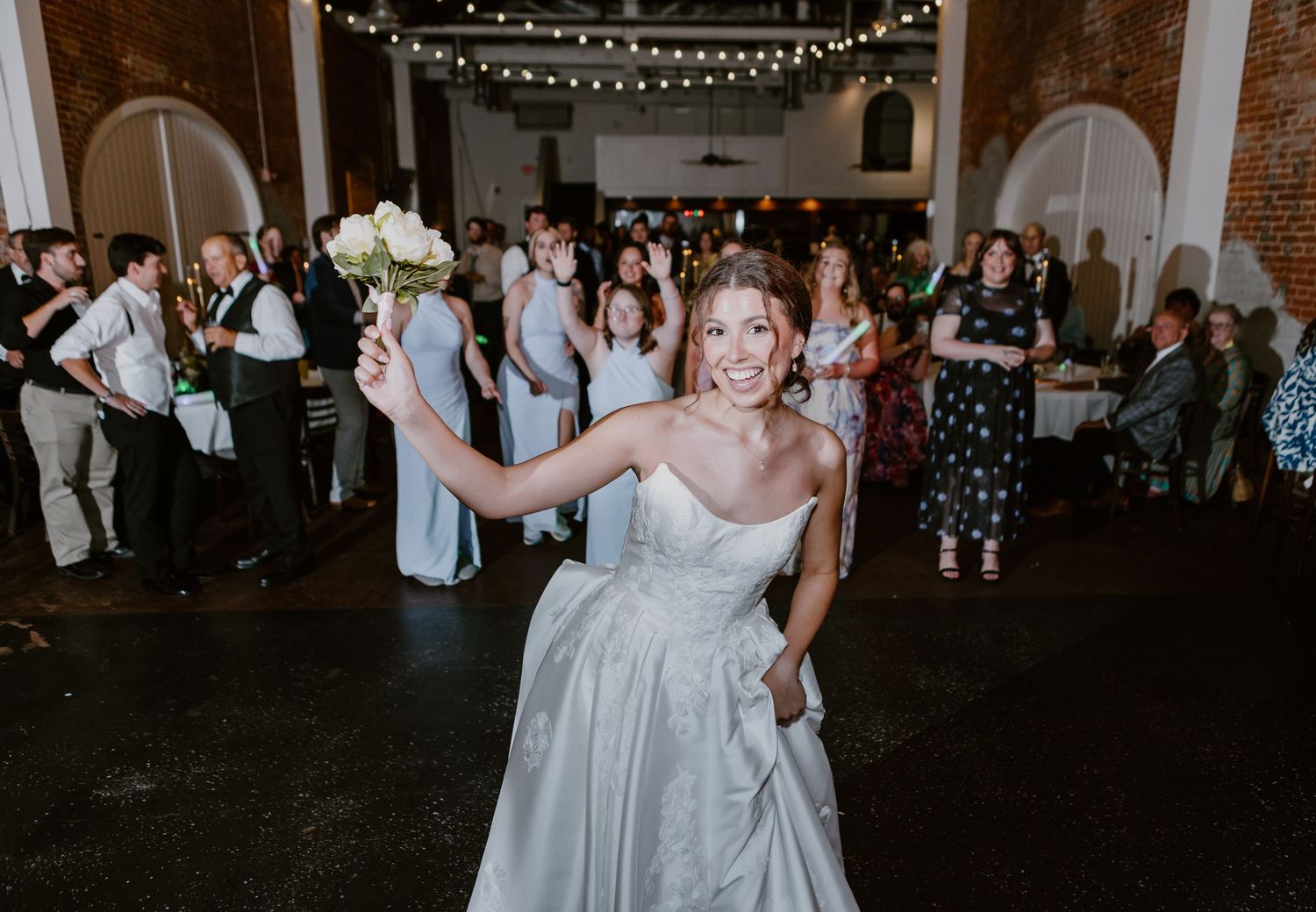 Bride in strapless gown tossing bouquet at indoor wedding reception with string lights and guests.