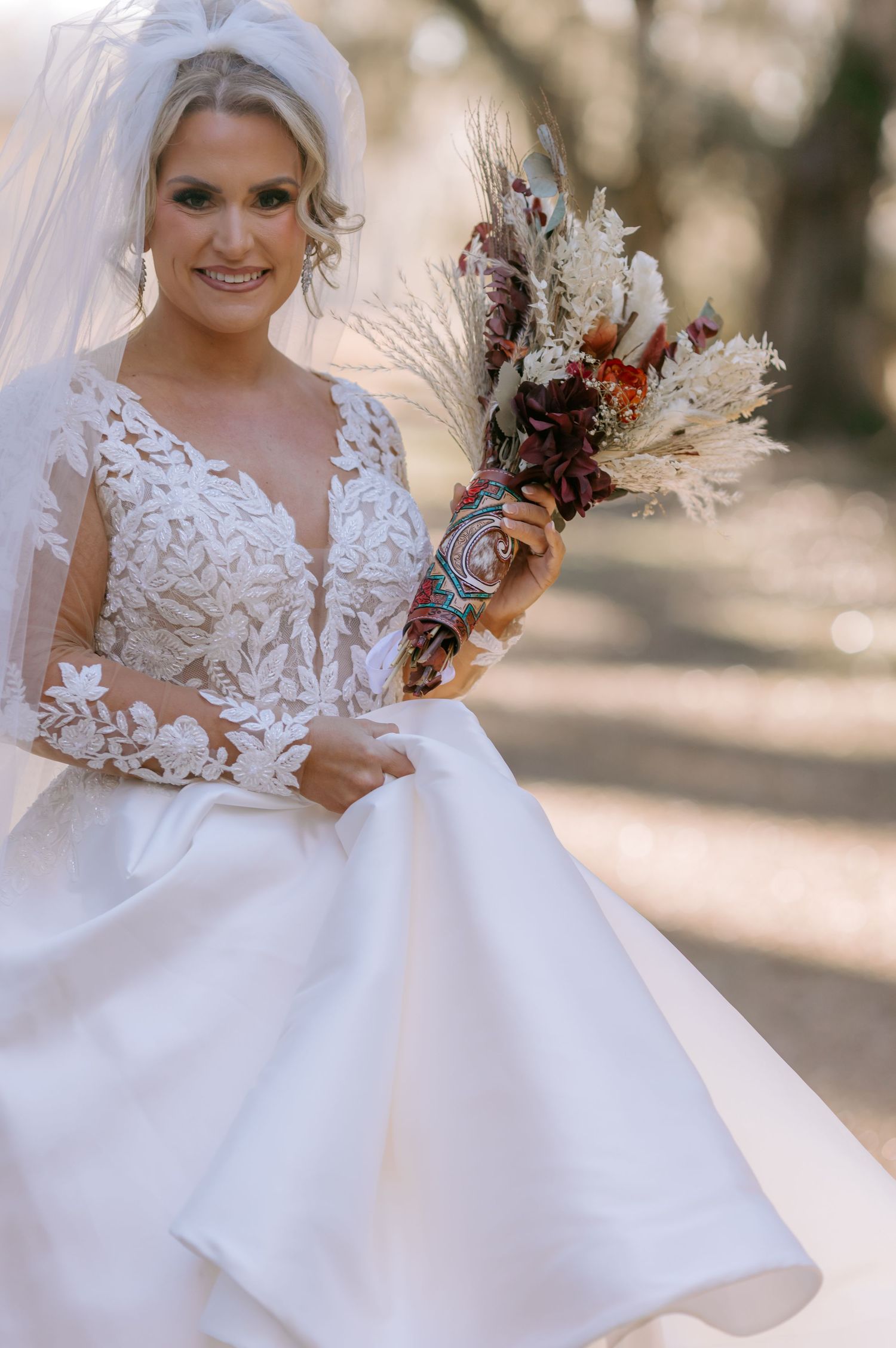 A bride in a white lace wedding dress holds a stunning dried floral bouquet with pampas grass and burgundy accents.