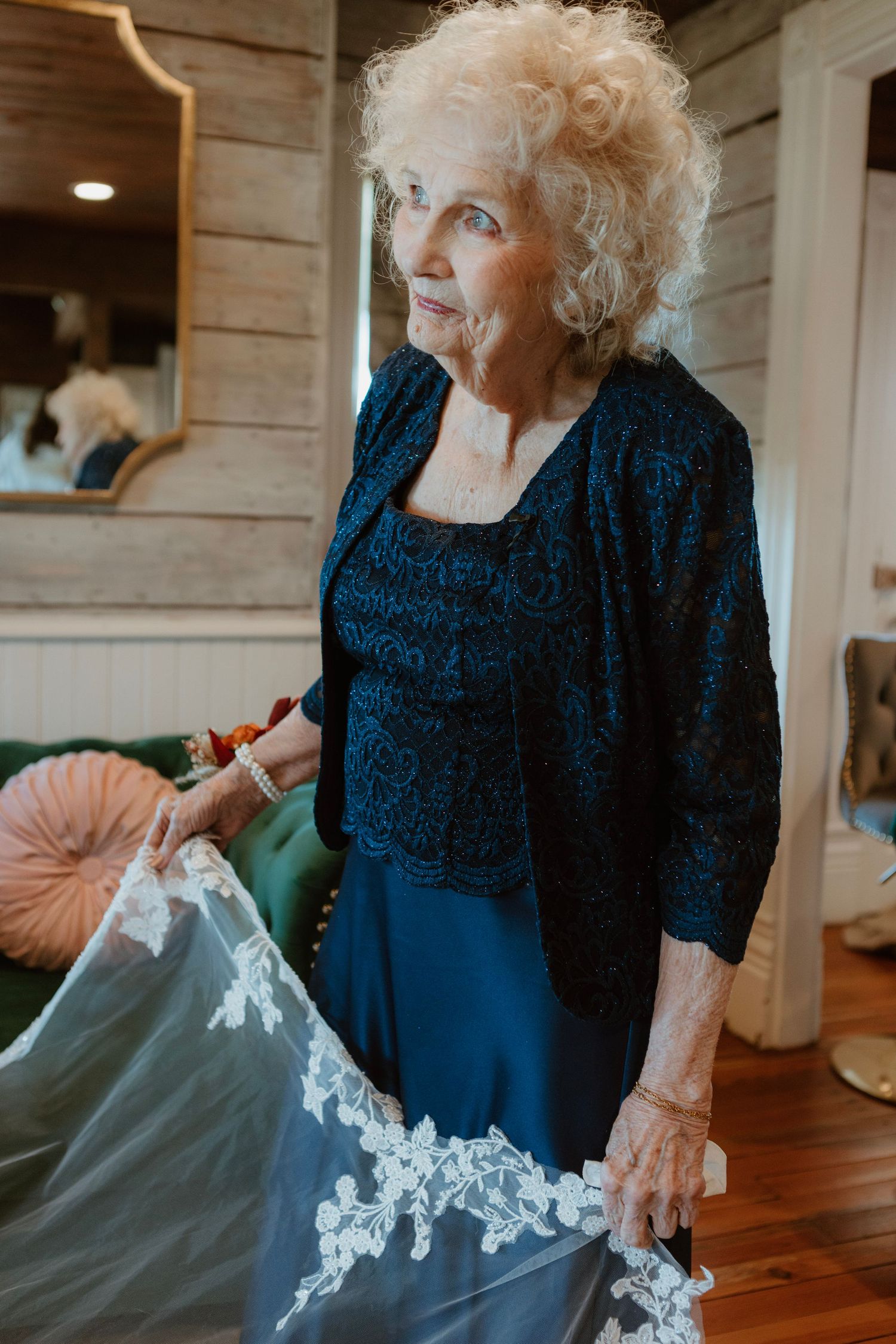 Elegant elderly woman in navy blue lace dress holding a white wedding gown with lace trim in a rustic interior.