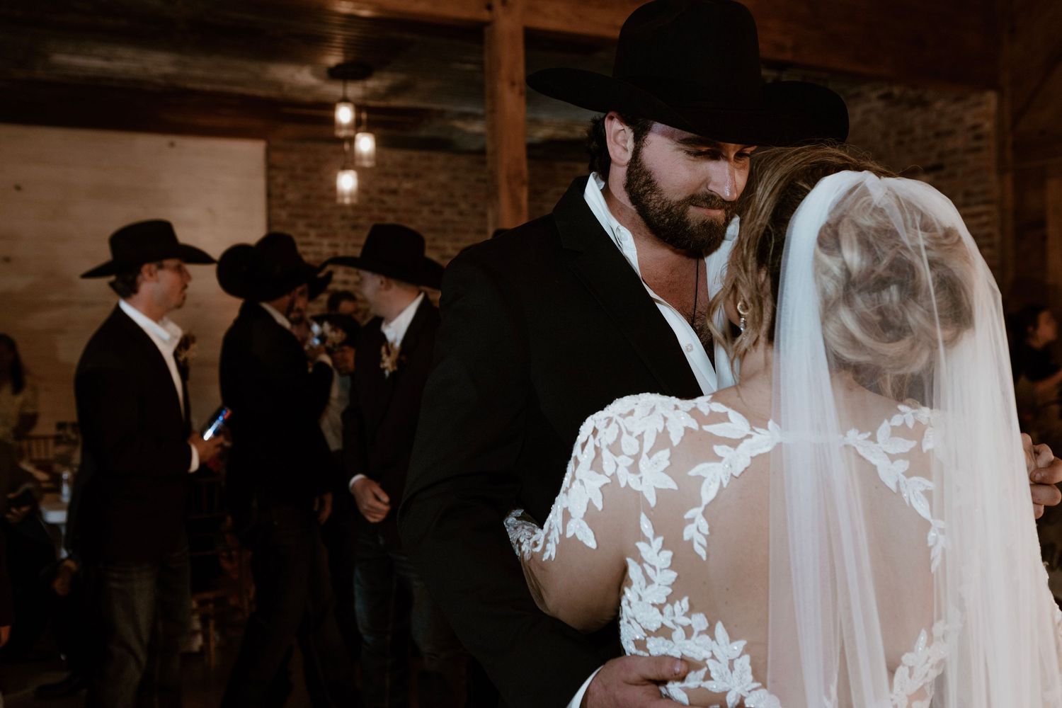 Bride and groom share first dance in rustic barn venue, groom in black cowboy hat, bride in lace wedding gown with veil.