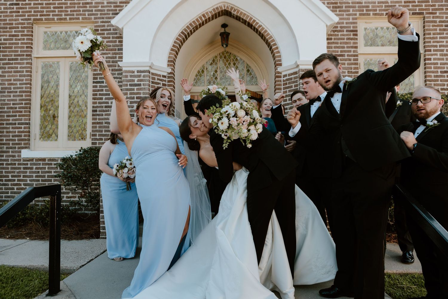 Joyful wedding party celebrating outside a brick church, bridesmaids in blue dresses and groomsmen in black tuxedos.