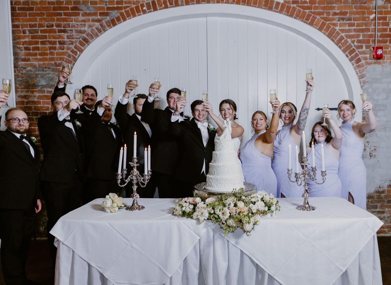 Wedding party toasting with champagne around a white cake table with candelabras in a brick venue.