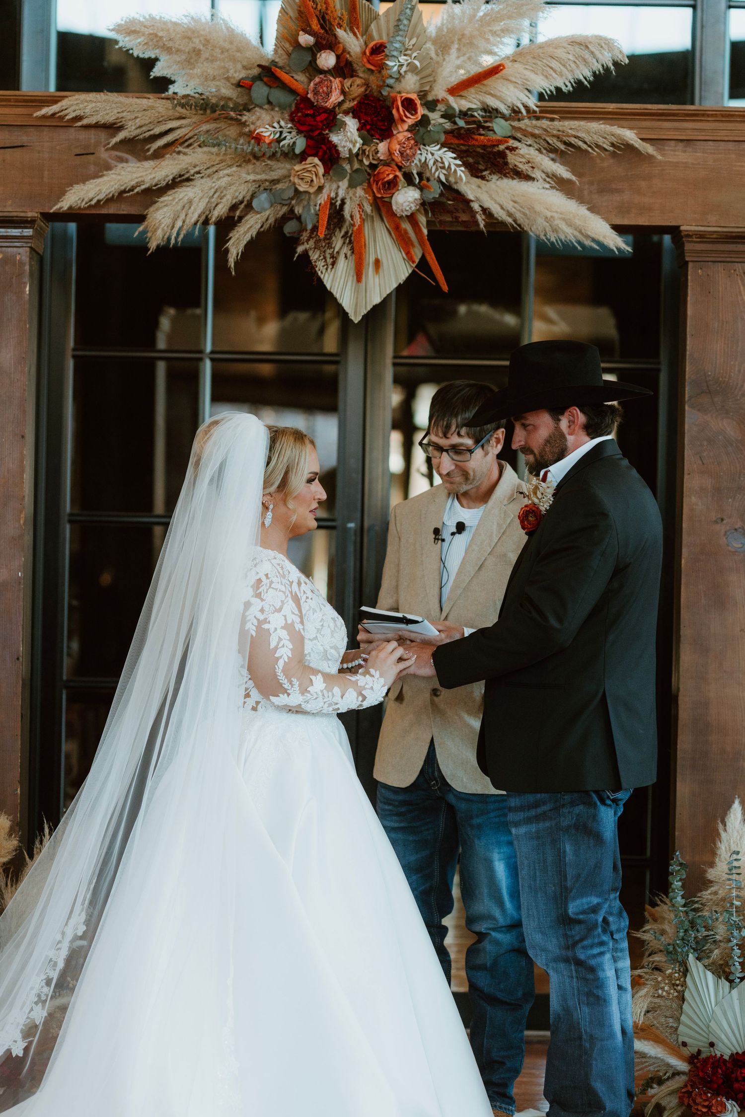 Bride and groom exchange vows at rustic wedding ceremony with pampas grass floral arch and officiant.