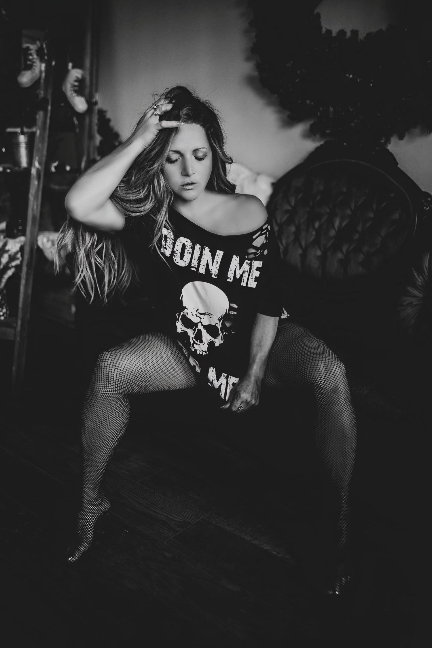 Dramatic black and white photo of a model in fishnet stockings and gothic shirt posing against dark background.