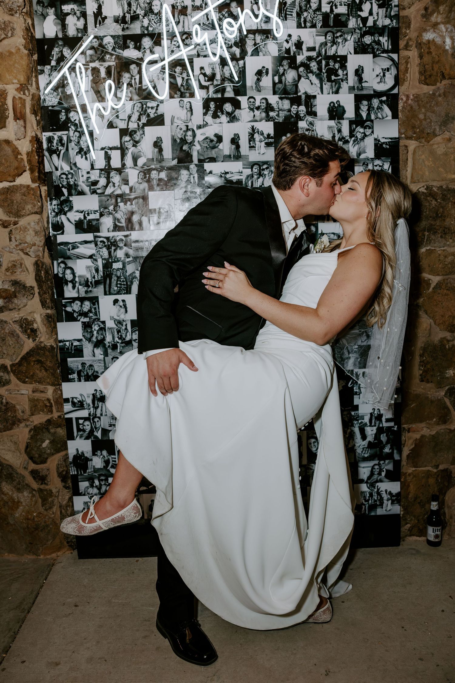 Bride in a white gown and veil is dipped in a romantic kiss by the groom in a black suit in front of a black-and-white photo wall.