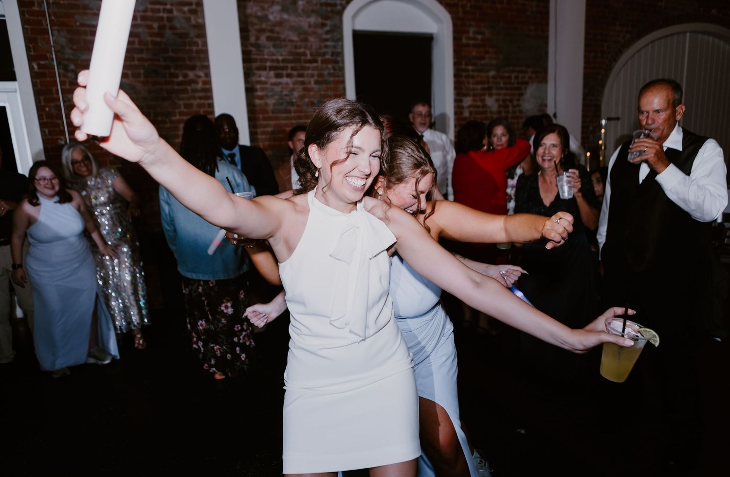 Bride in white mini dress dancing joyfully at wedding reception with guests celebrating in background.