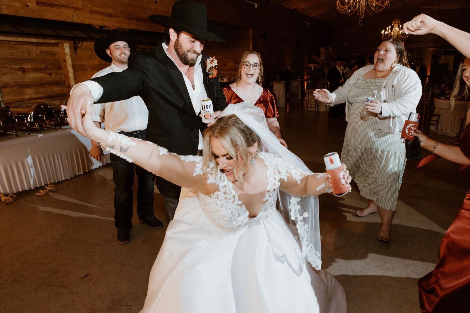 Bride in lace gown dances joyfully with groom in cowboy hat at reception.