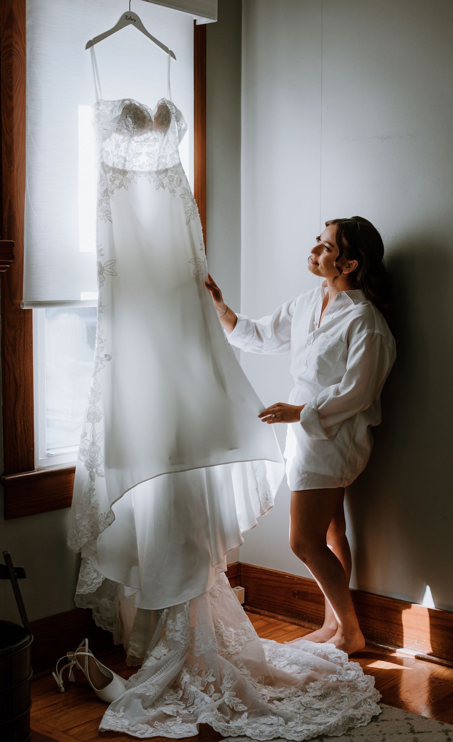 Bride in white robe admiring her lace wedding gown hanging by a sunlit window before the ceremony.