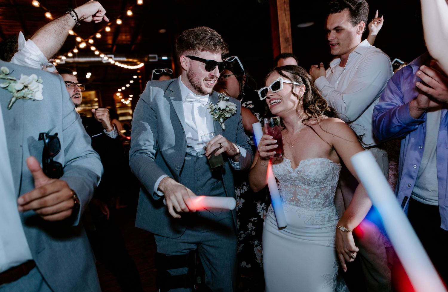 Bride and groom wearing white sunglasses celebrate with drinks and glow sticks on the dance floor.