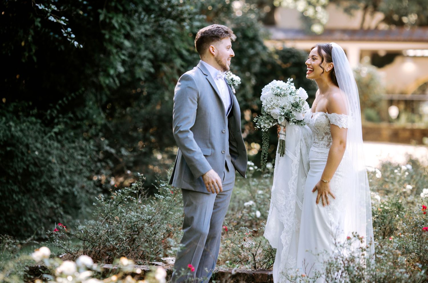Smiling bride in off-shoulder lace gown and groom in gray suit share a joyful moment in a rose garden.