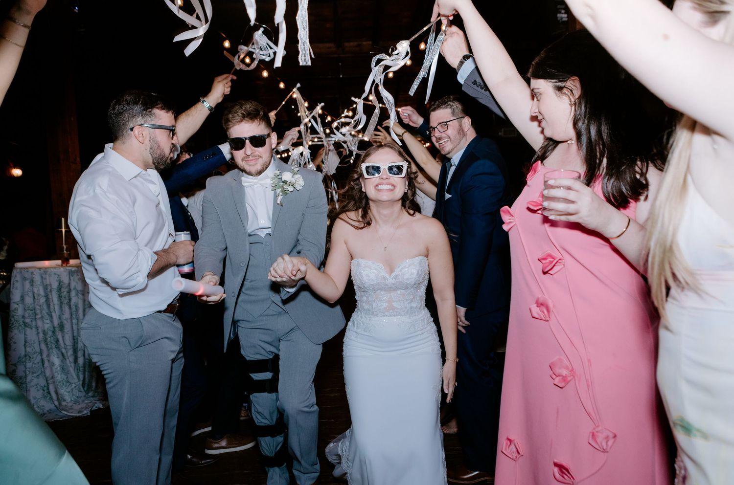 Bride and groom in sunglasses celebrate wedding reception exit with cheering guests in elegant venue.