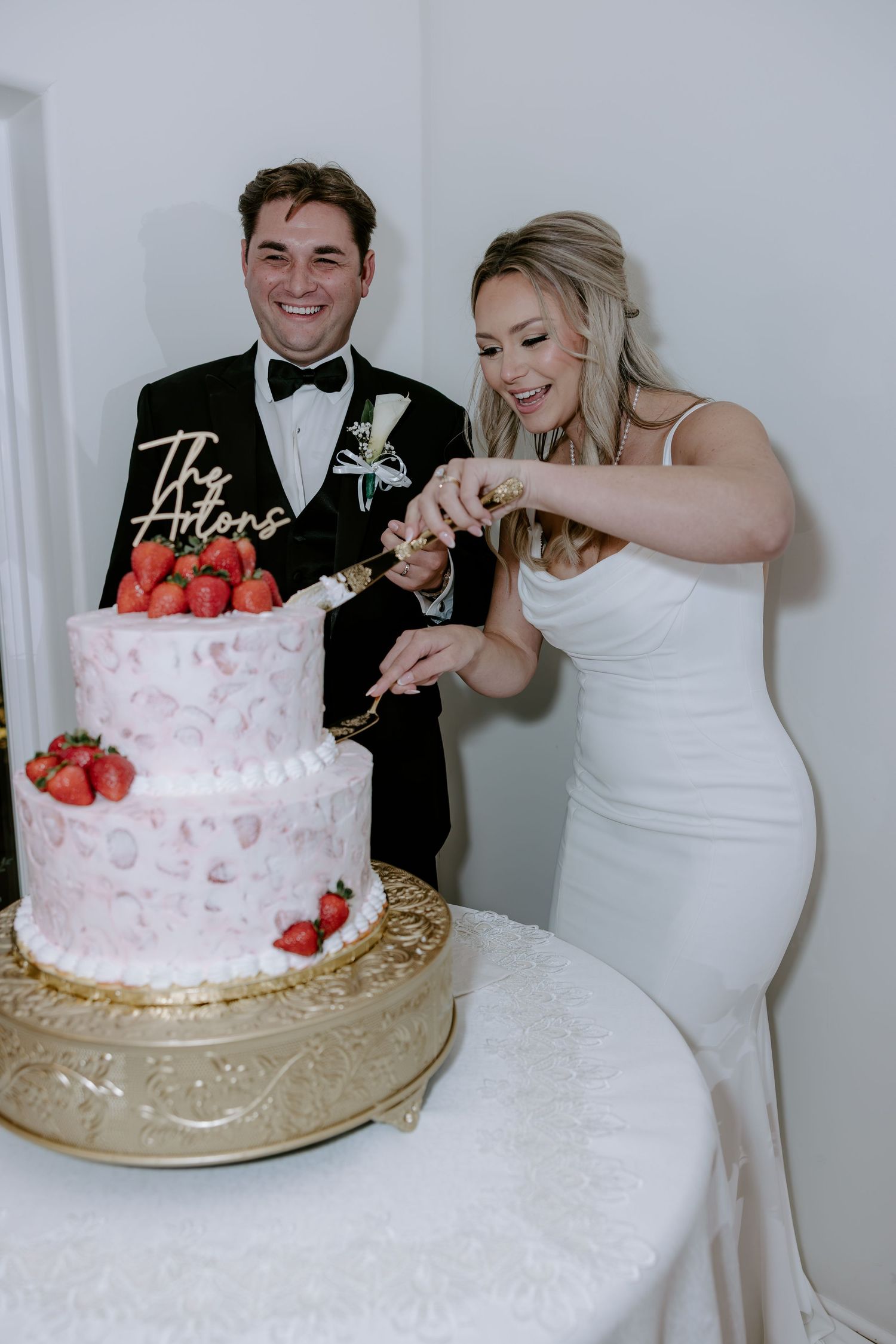 A smiling bride in a white gown and groom in a black tuxedo cut their pink marbled wedding cake topped with fresh strawberries.
