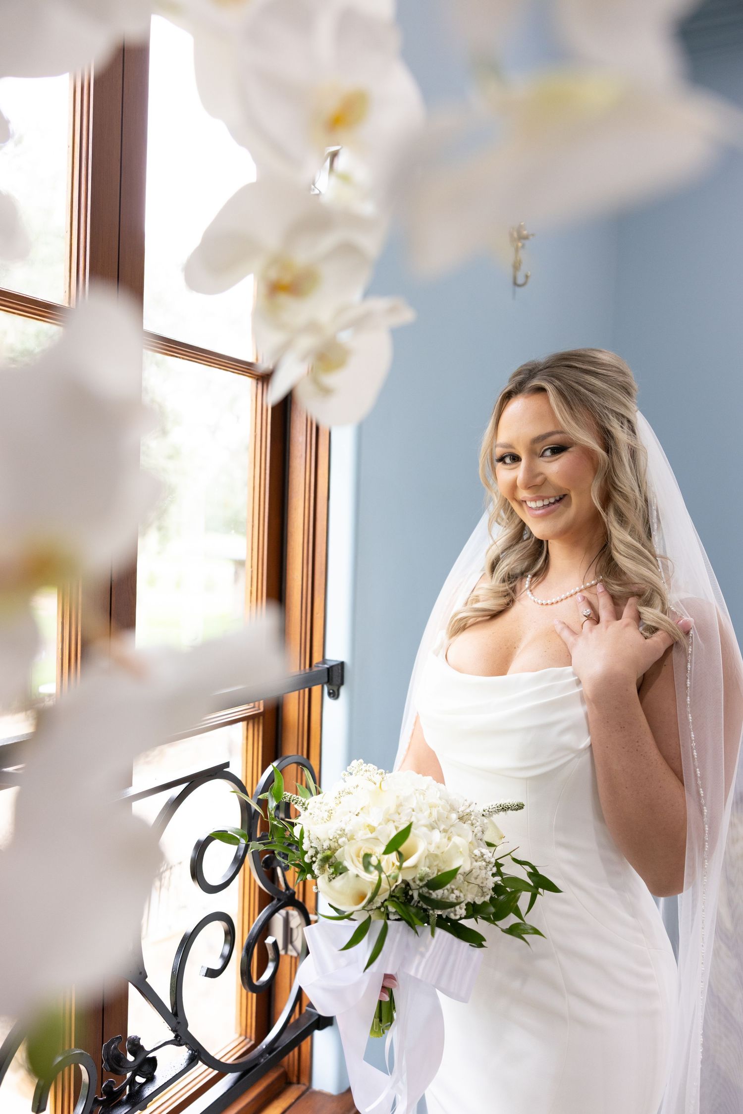 Smiling bride in a white satin wedding gown and long veil holds a white floral bouquet beside a window with ornate iron railing.