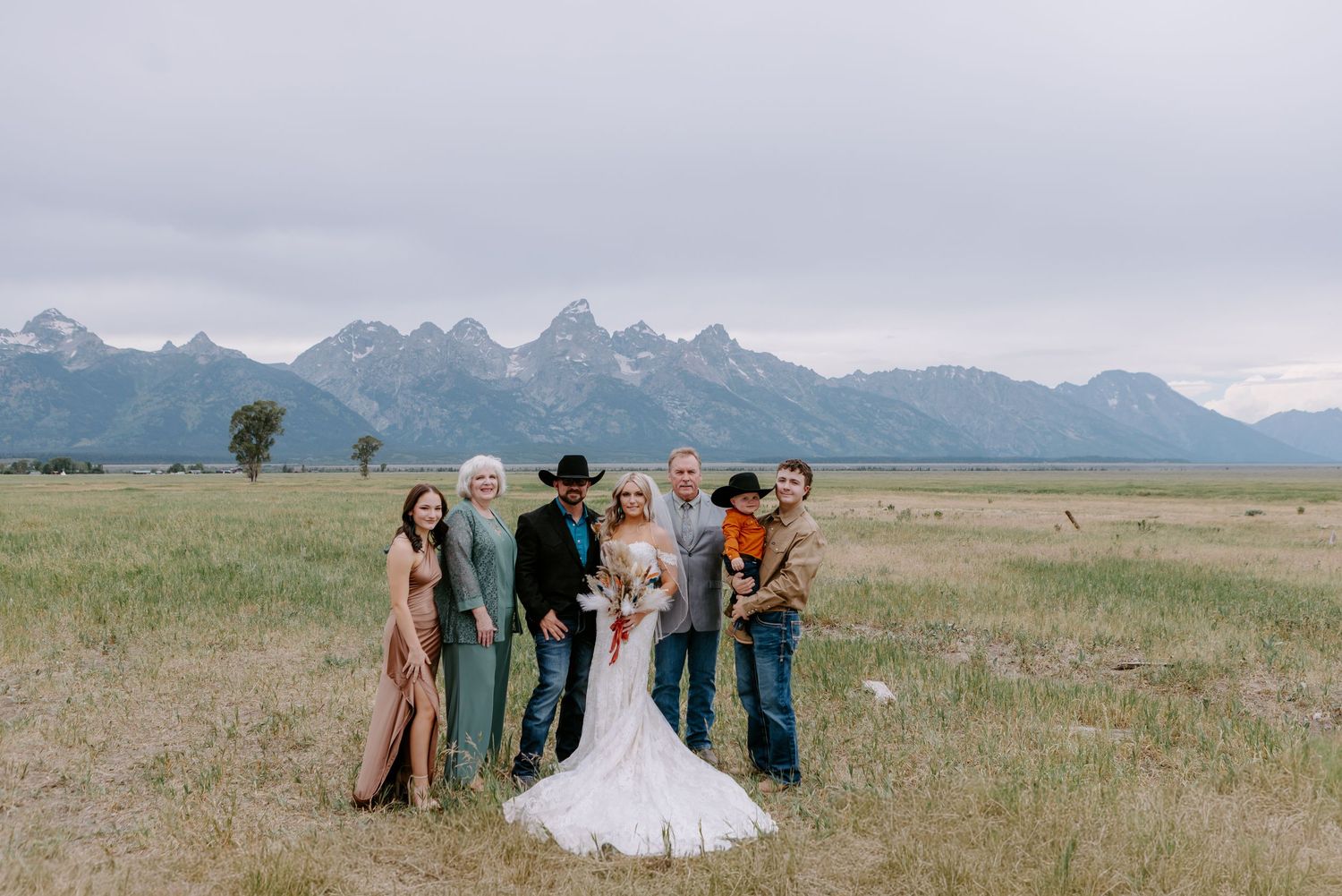 Wedding party poses in a lush green field with the Grand Teton mountain range in the background on a cloudy day.