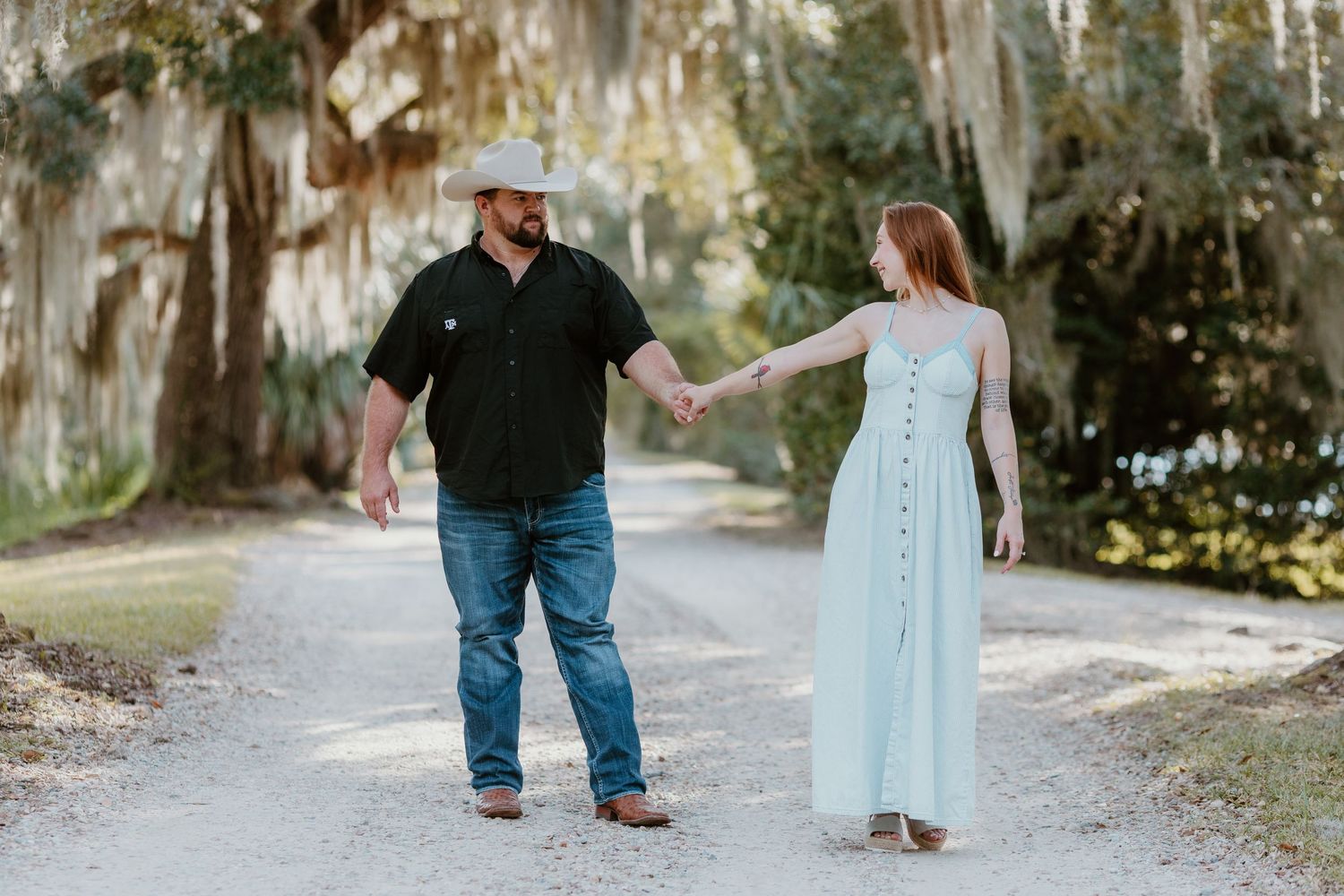 A couple walking together on a sunlit gravel path surrounded by hanging Spanish moss and trees.