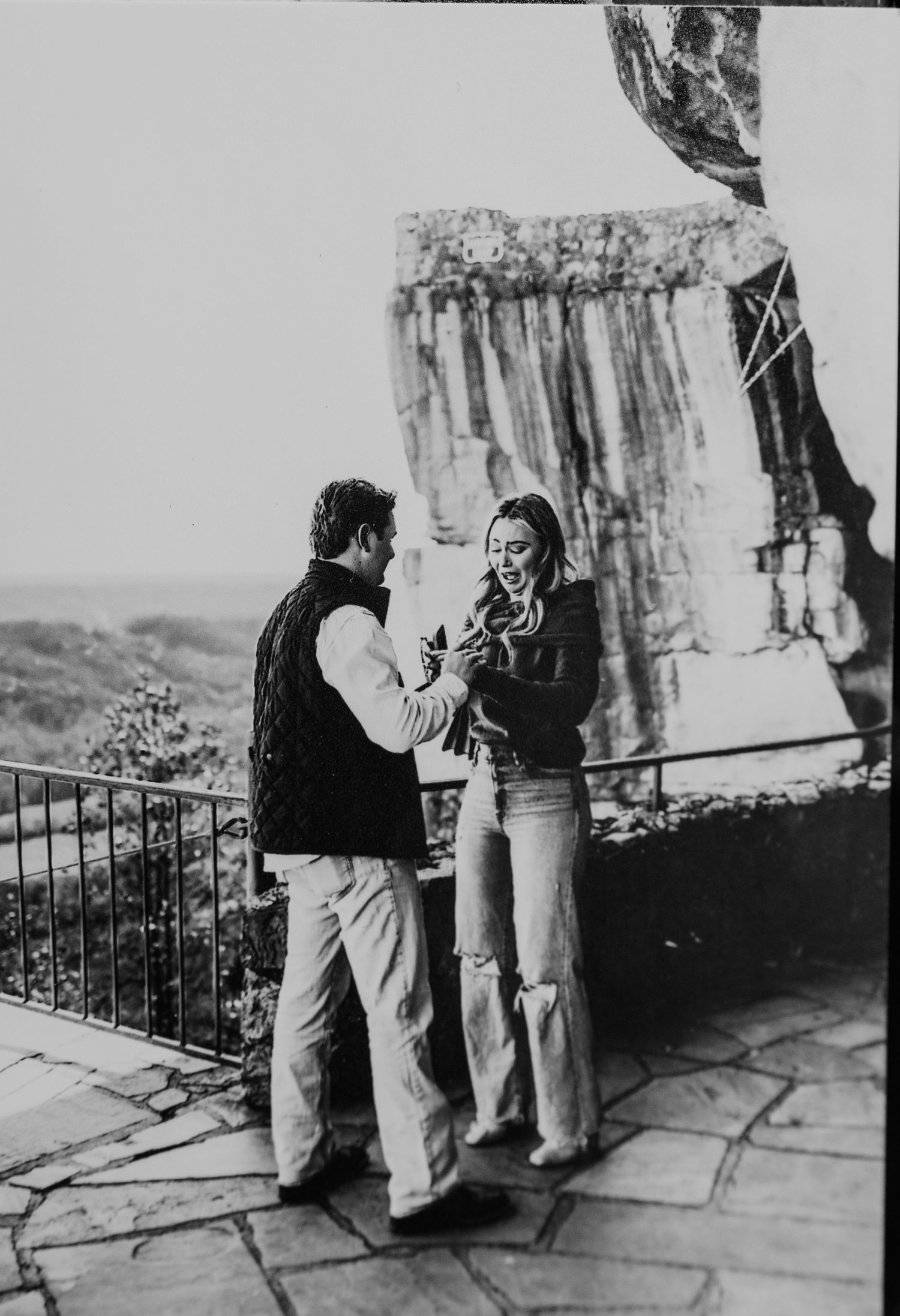 Black and white photo of a couple sharing an intimate moment on a stone overlook with a rock face and scenic valley behind them.