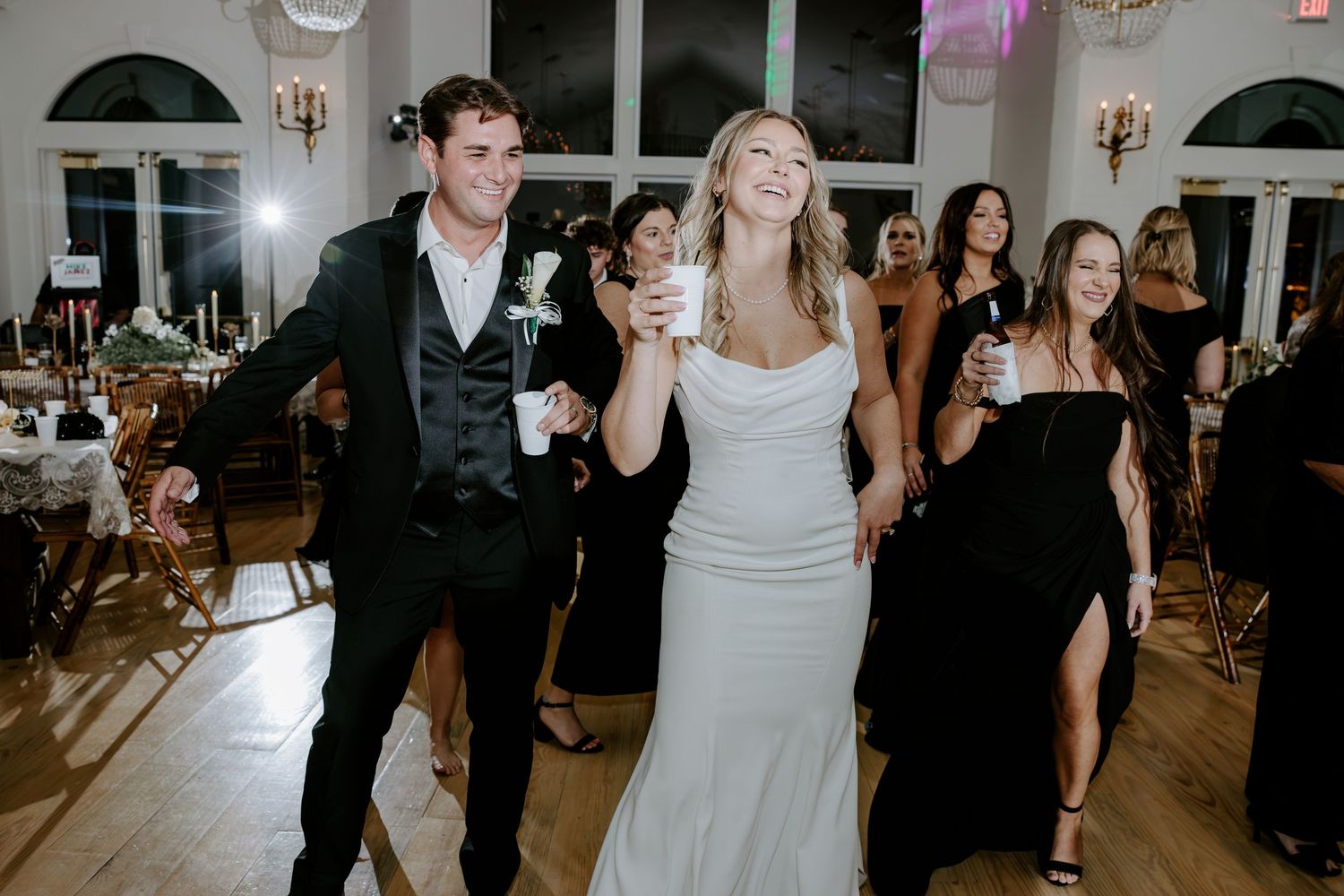 Bride in a white gown laughs and dances with a groom in a black vest as wedding guests in black dresses celebrate in an elegant ballroom.