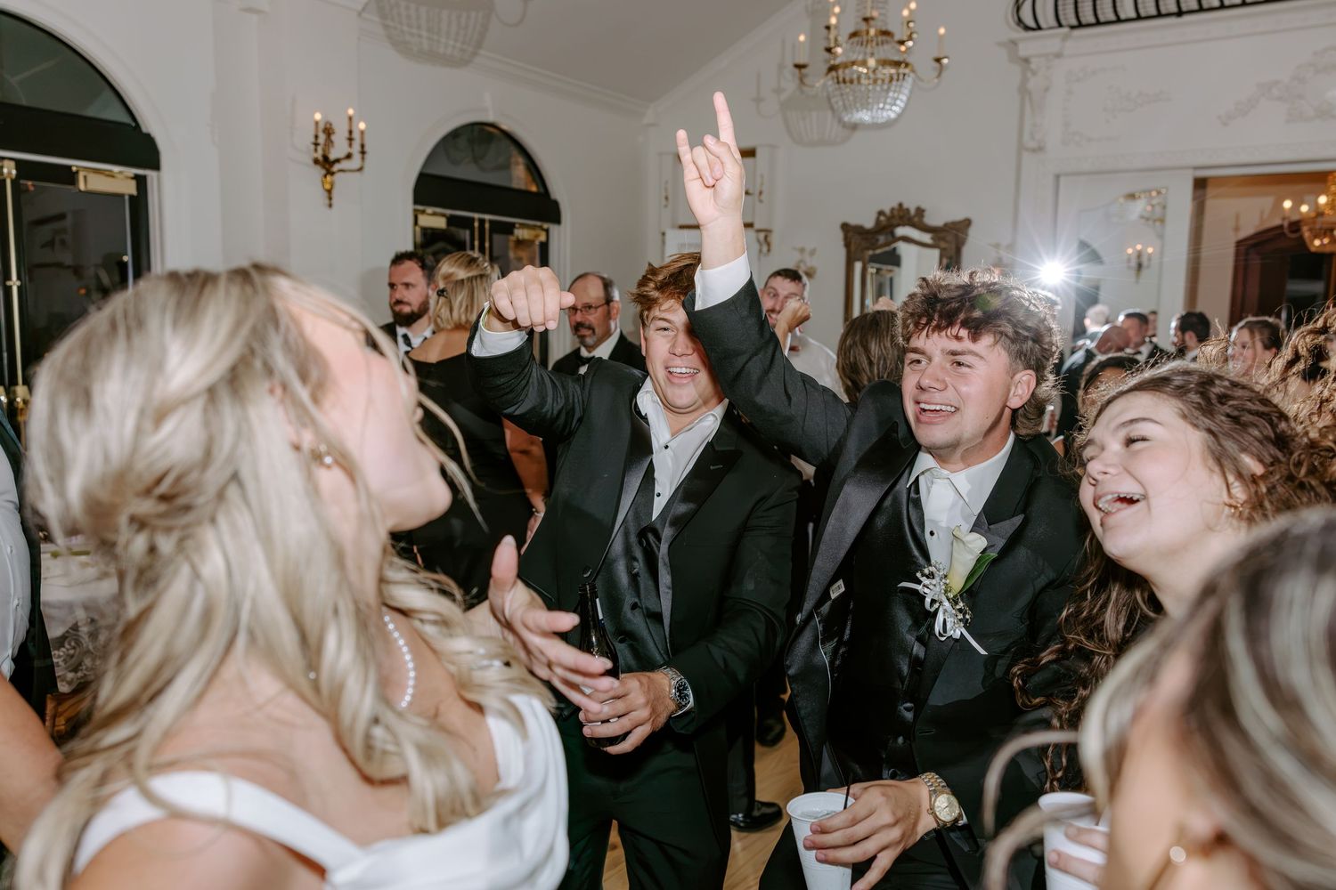 Wedding guests in formal attire dance and cheer with raised hands at a lively reception inside an elegant ballroom with chandeliers.