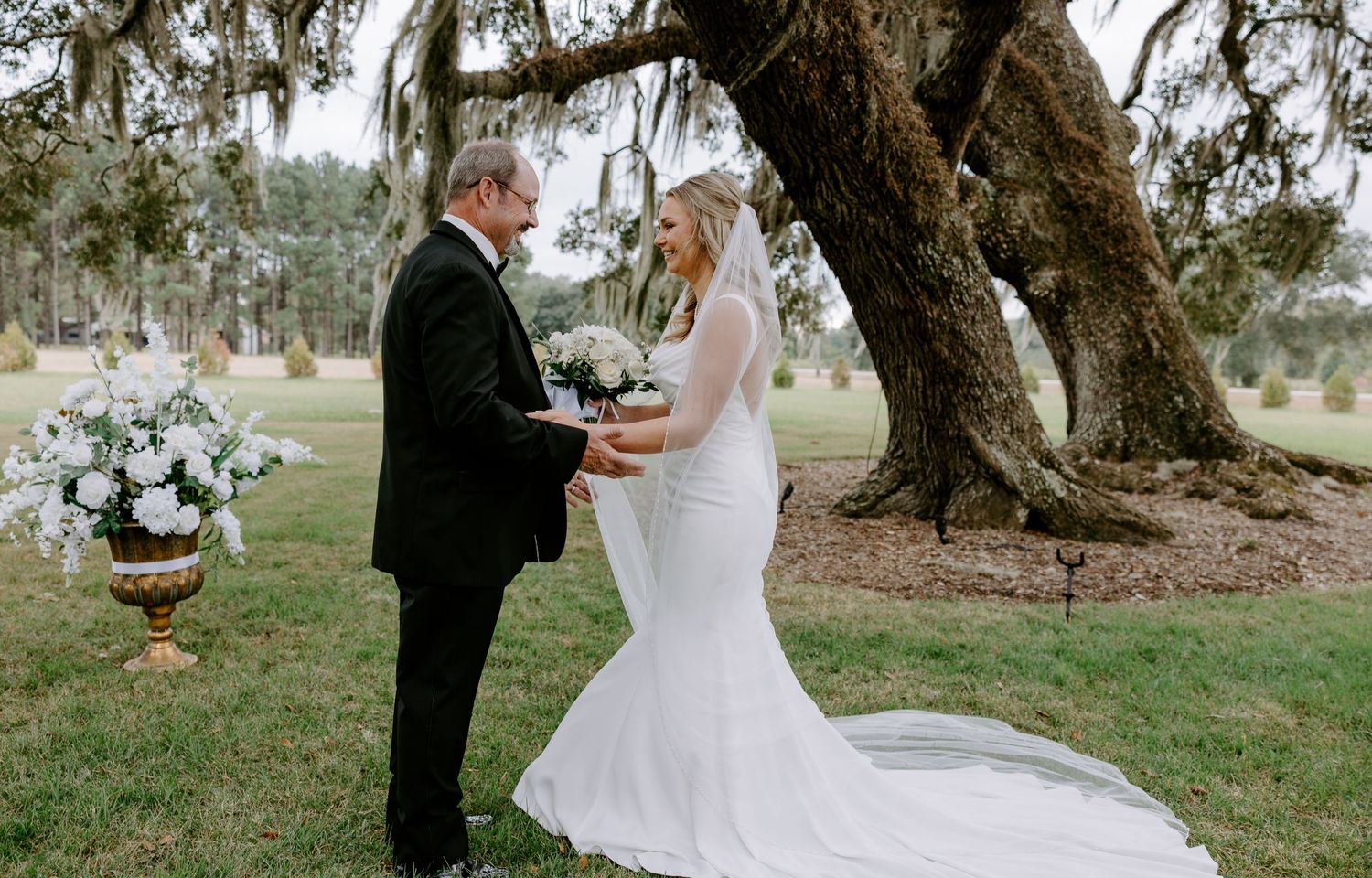 Bride in a white gown and groom in a dark suit hold hands under a live oak tree draped in moss at an outdoor wedding ceremony.