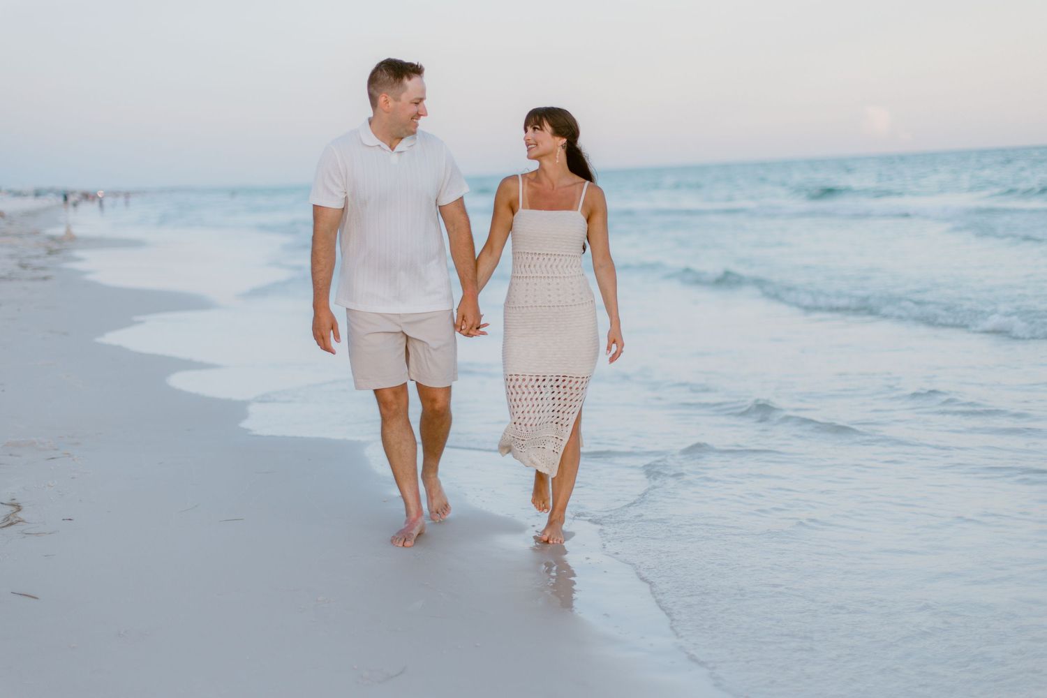 Couple walking hand in hand along a white sandy beach at sunset with gentle waves rolling in.
