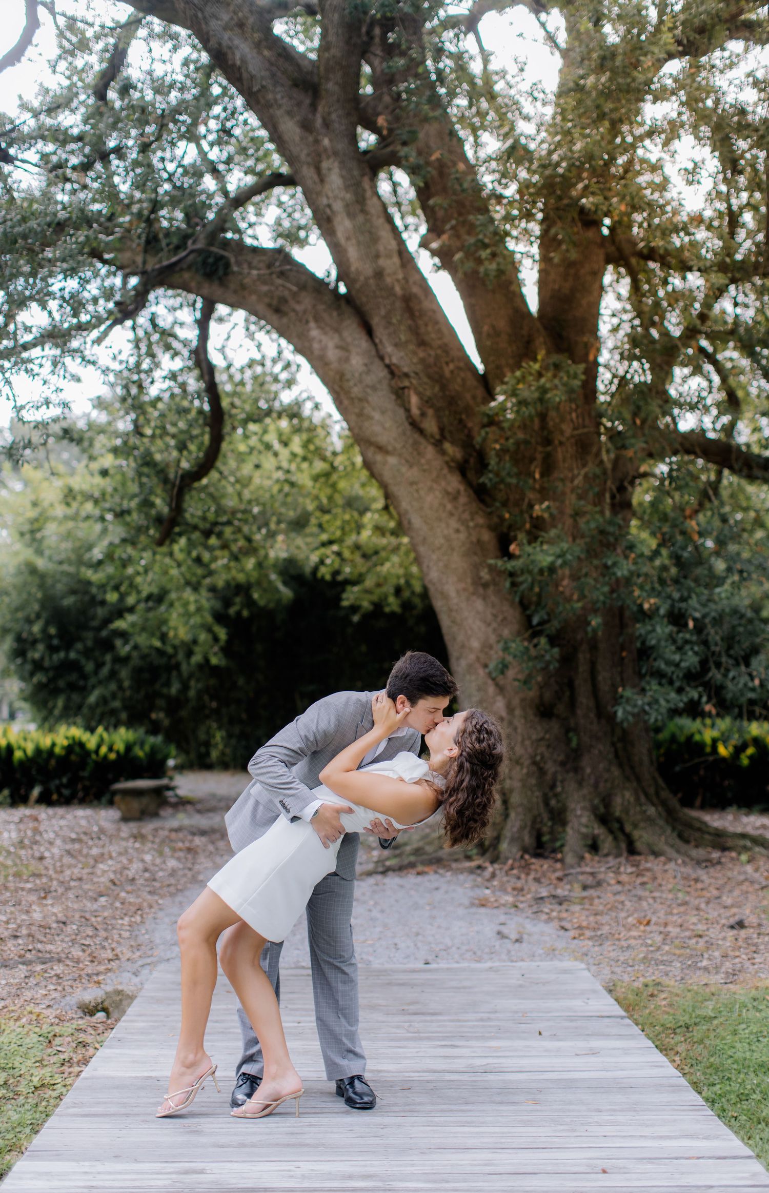 A couple performs an elegant dip dance move on a wooden walkway surrounded by lush trees and natural scenery.
