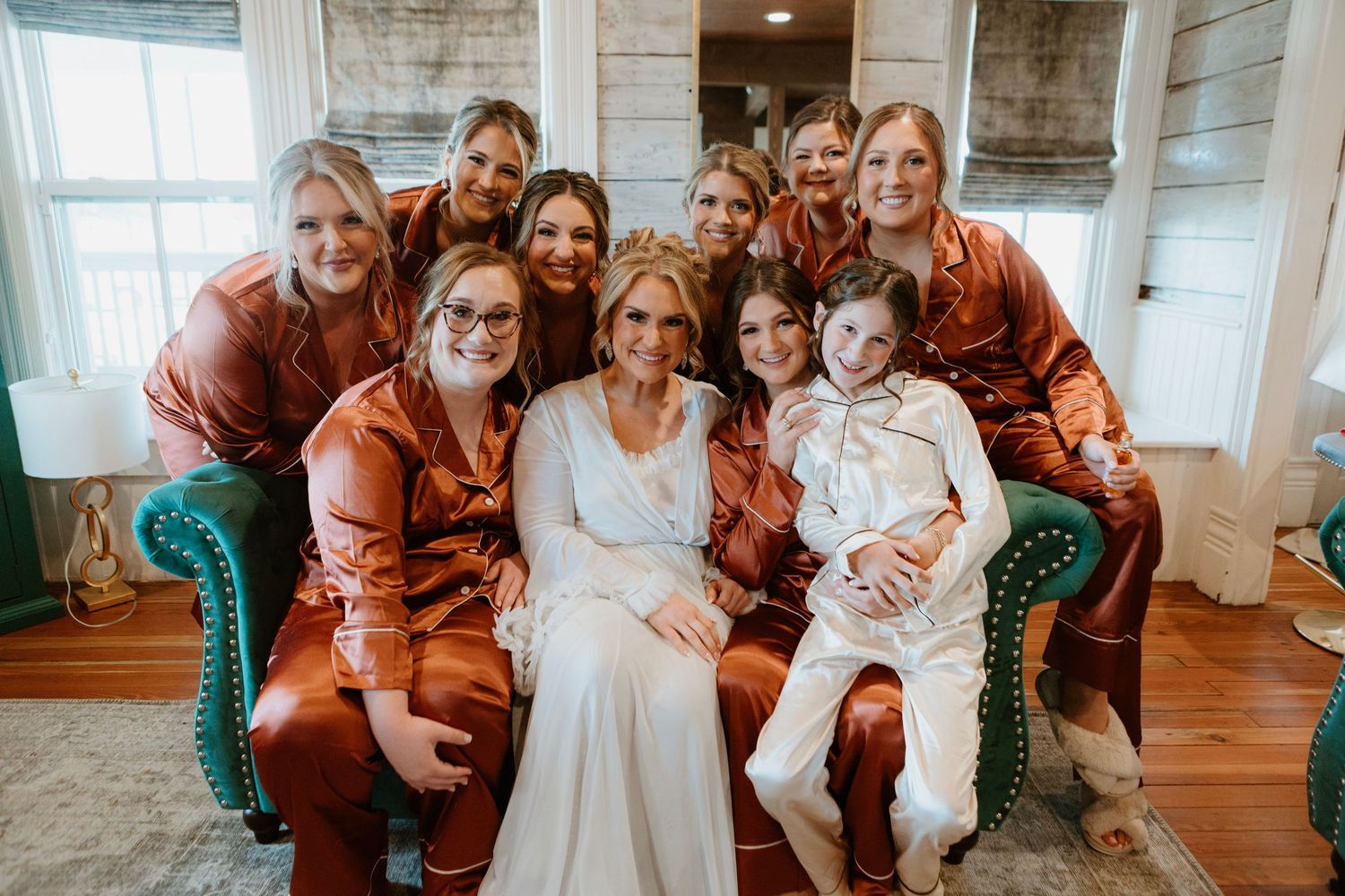 Bride in white gown poses with bridesmaids in rust satin robes and a child on a teal chair indoors.