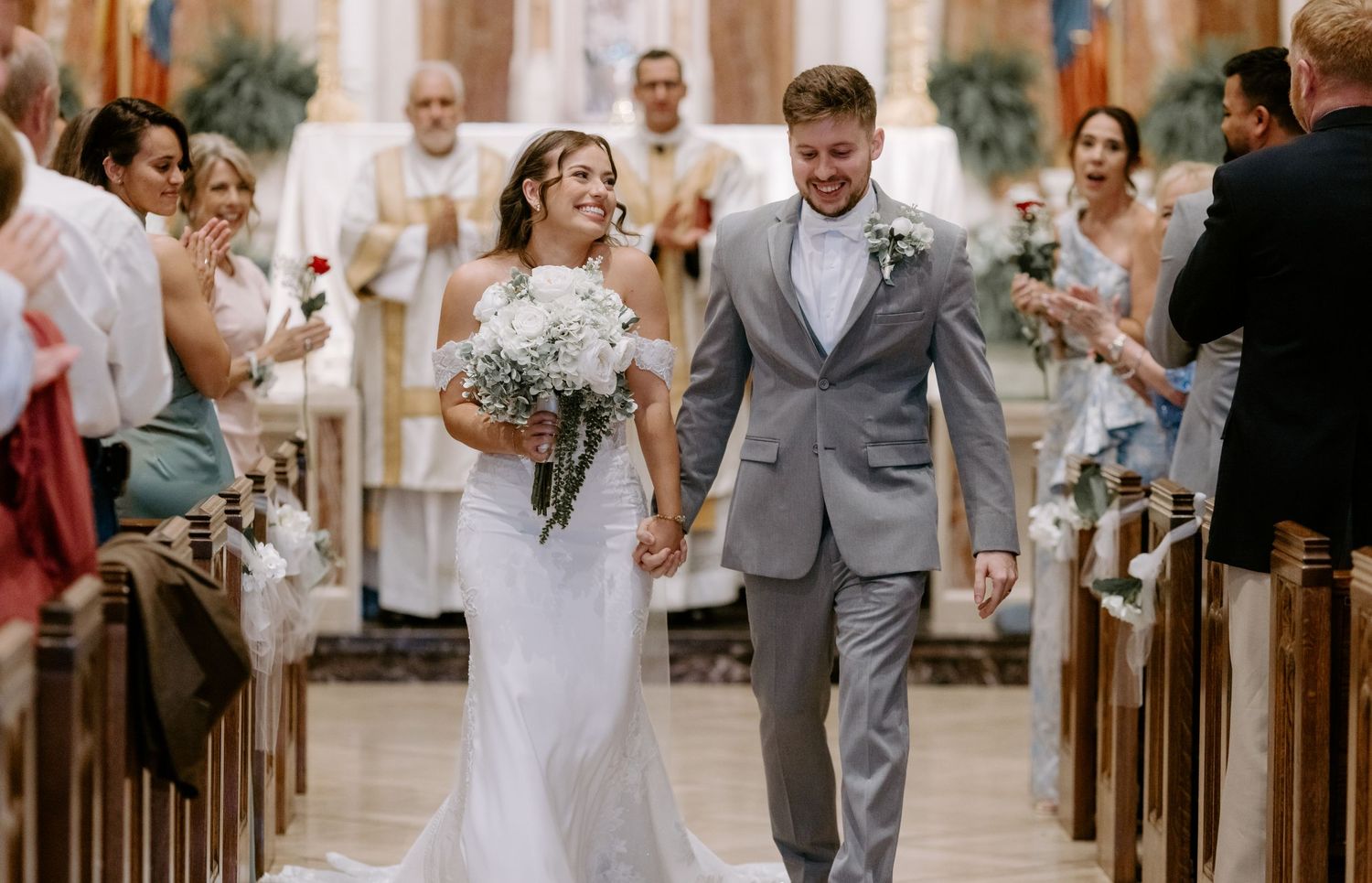 Bride and groom walk hand-in-hand down church aisle after wedding ceremony, guests celebrating around them.