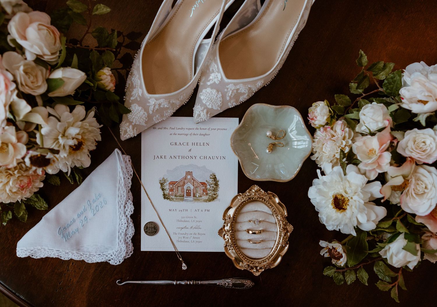 Elegant wedding flat lay featuring lace heels, floral bouquets, invitation, rings in gold frame, and pearl accessories on wood.
