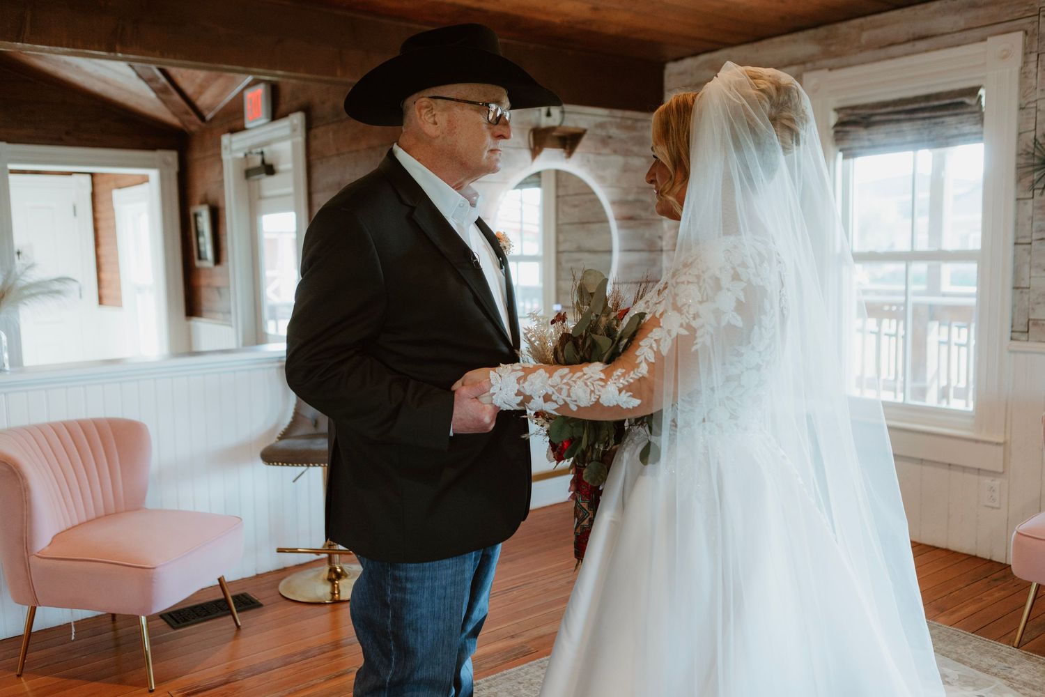 Father seeing bride in wedding dress for first time, holding hands in rustic venue.