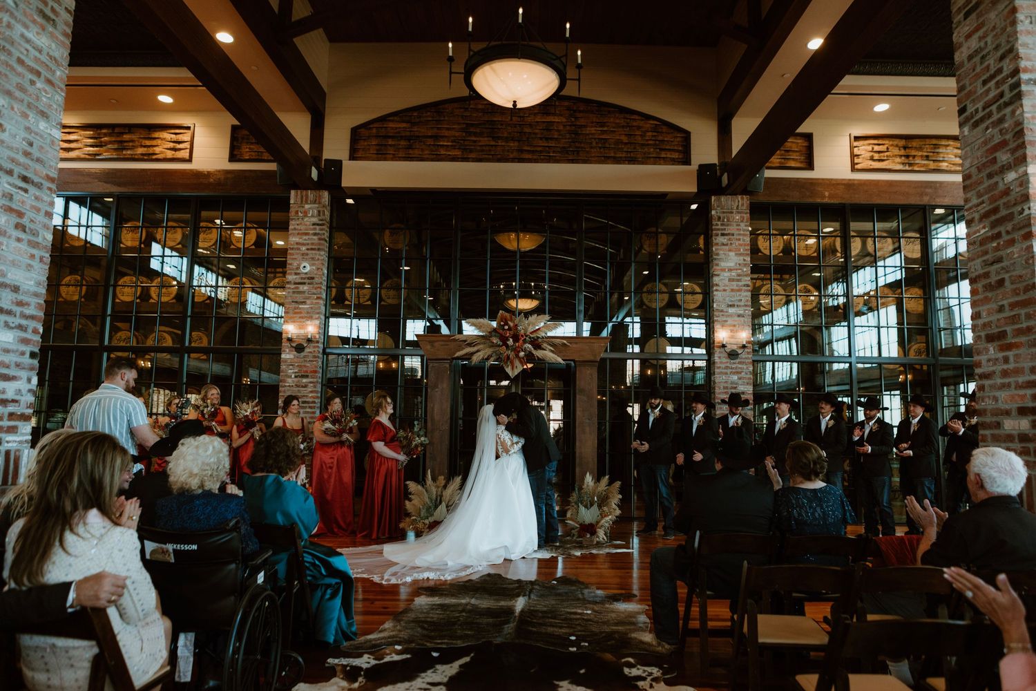 Elegant indoor wedding ceremony in a rustic venue with brick columns, large windows, and guests seated in rows.