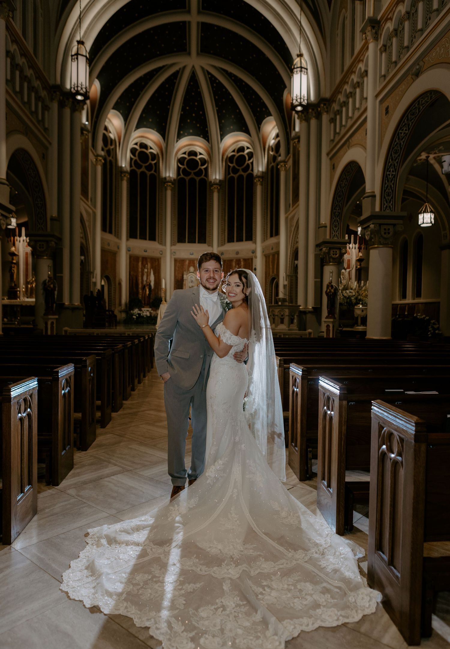 Bride and groom pose together in an elegant church aisle with arched ceilings, wooden pews, and a long lace wedding veil.