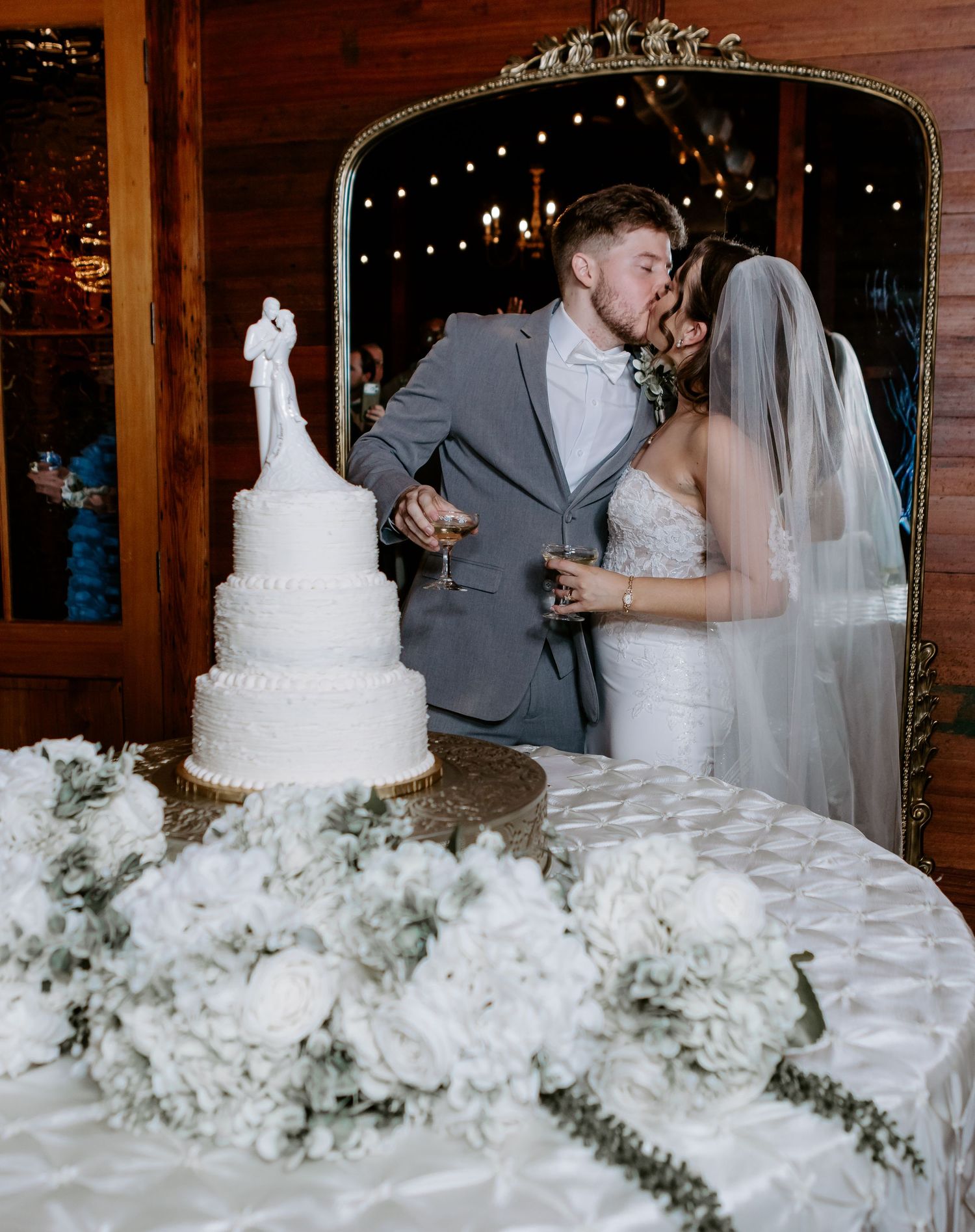 Bride and groom share a kiss behind their elegant white wedding cake adorned with floral arrangements at their reception.