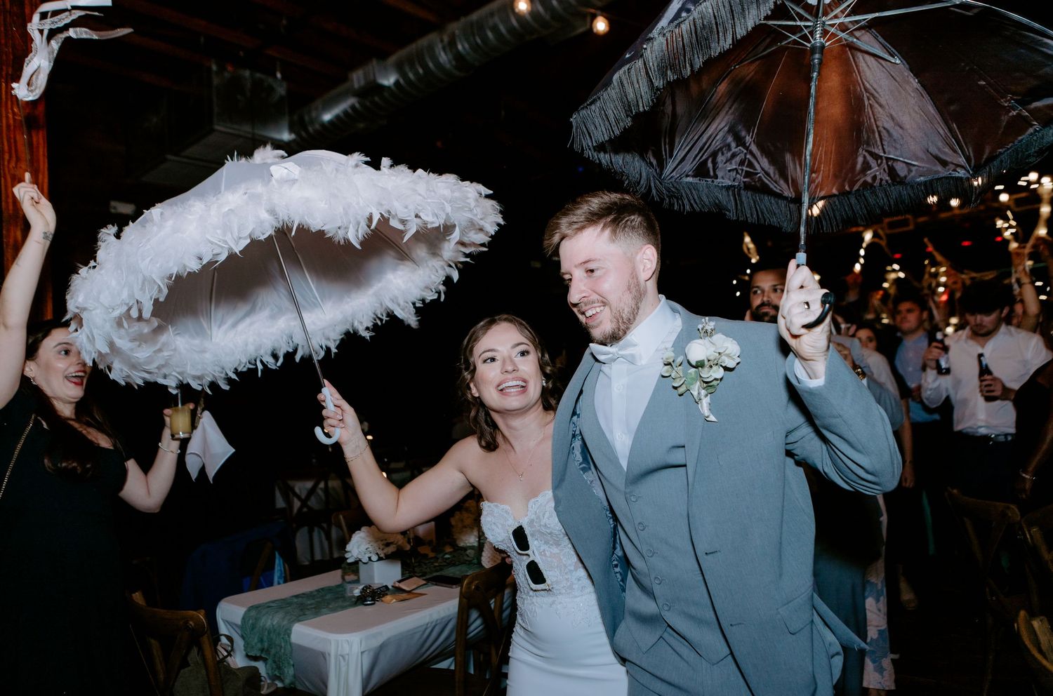 Bride and groom celebrate with feathered umbrellas during festive wedding reception exit.