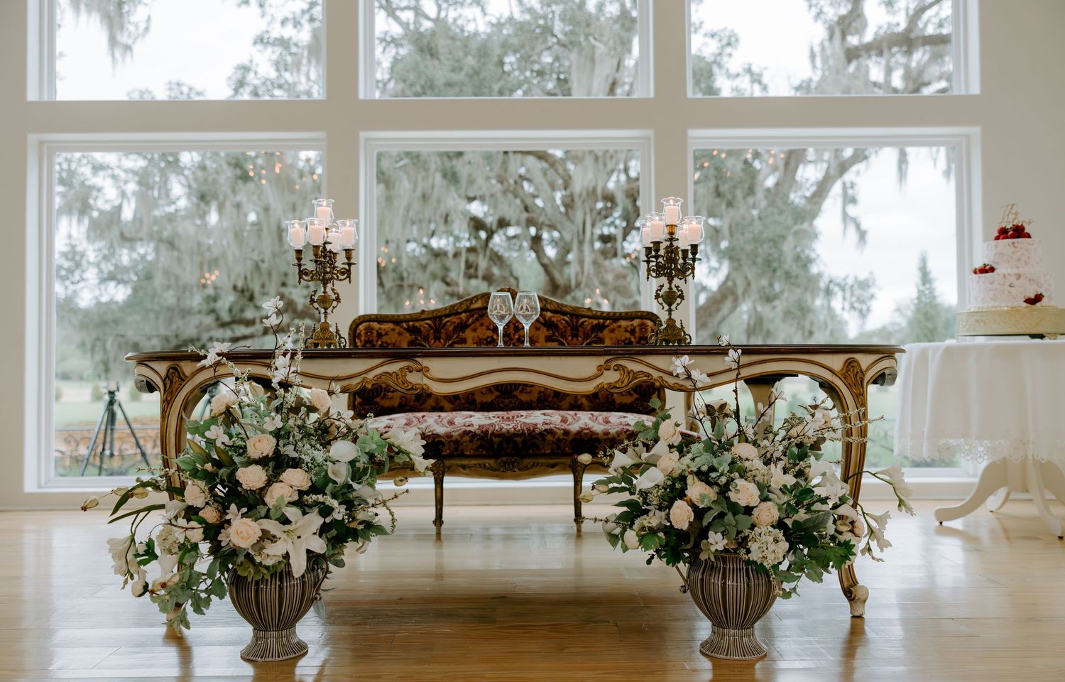 Ornate wedding venue interior with an antique gold bench, white floral arrangements, candelabras, and tall windows overlooking oak trees.