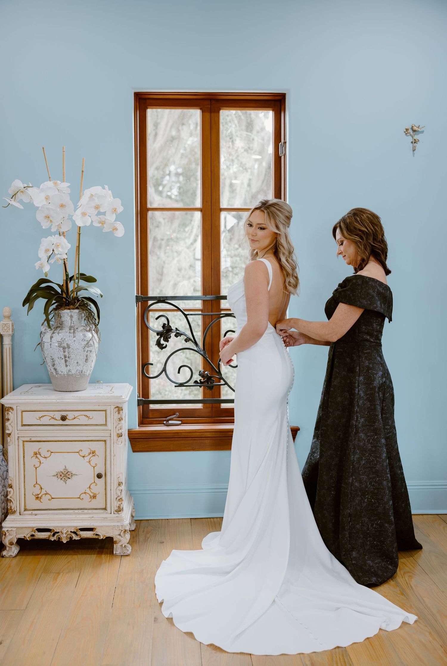 Bride in an elegant white gown with a long train has her dress buttoned by a woman in a black lace dress beside a window and white orchids.