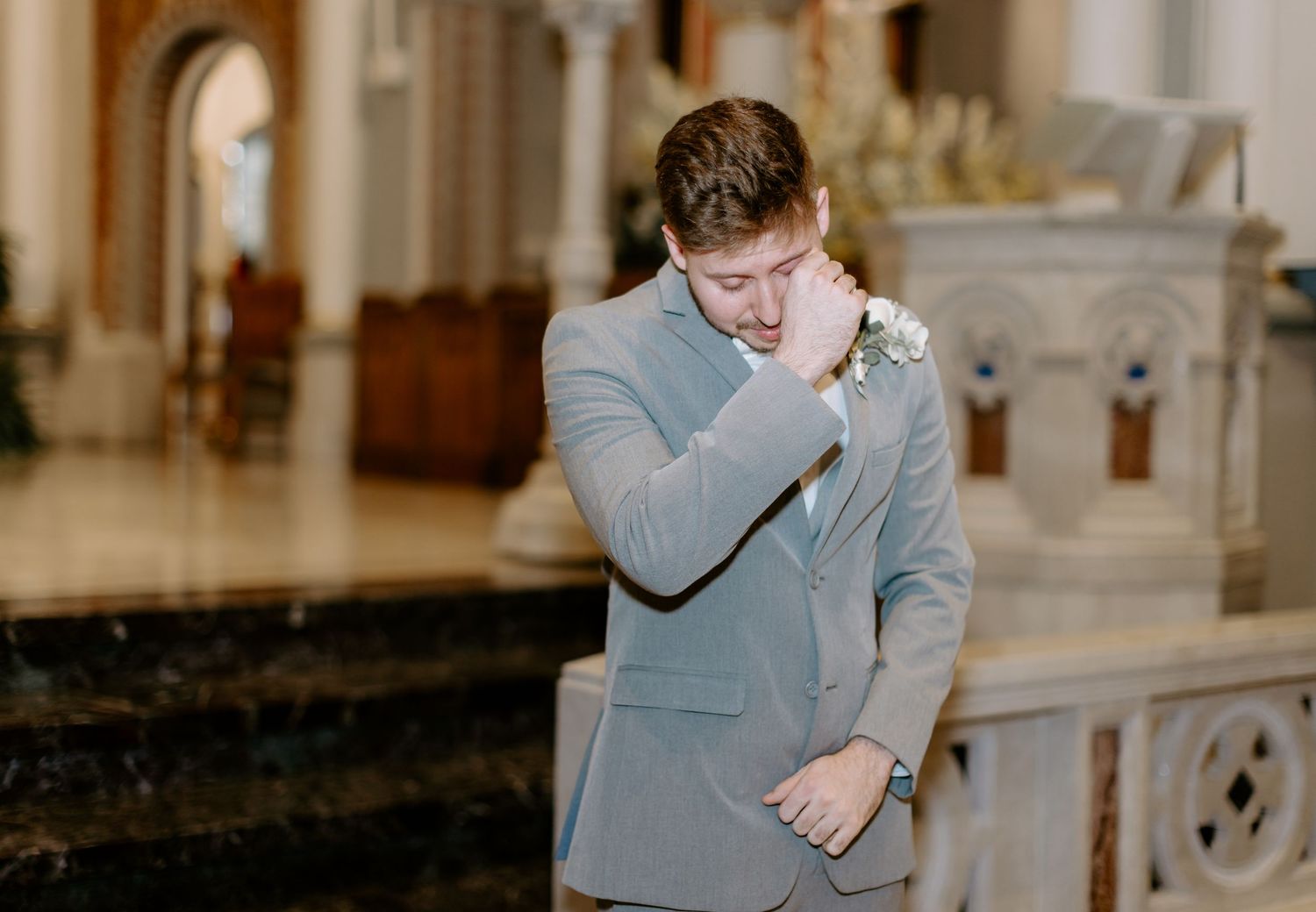 Emotional groom in gray suit wiping tears at church altar during wedding ceremony.