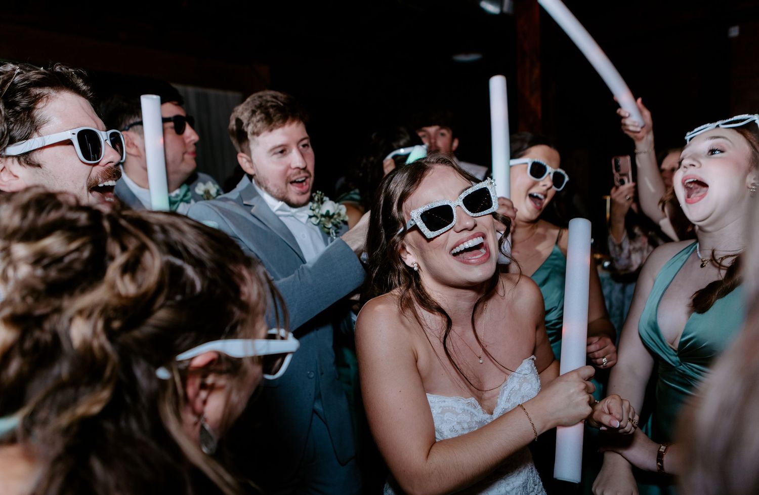 Bride and wedding guests dancing and laughing while wearing white sunglasses at reception party.