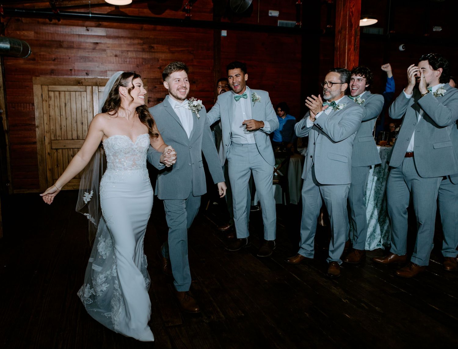 Bride in white lace gown and groom in gray suit make grand entrance at wedding reception with cheering groomsmen.