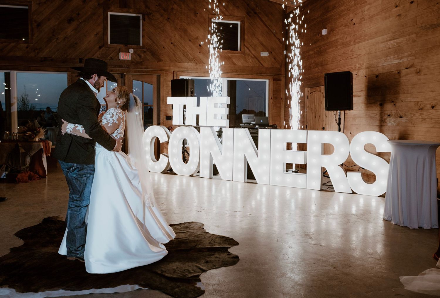Bride and groom share first dance in front of illuminated THE CONNERS marquee letters with sparklers at rustic barn wedding.