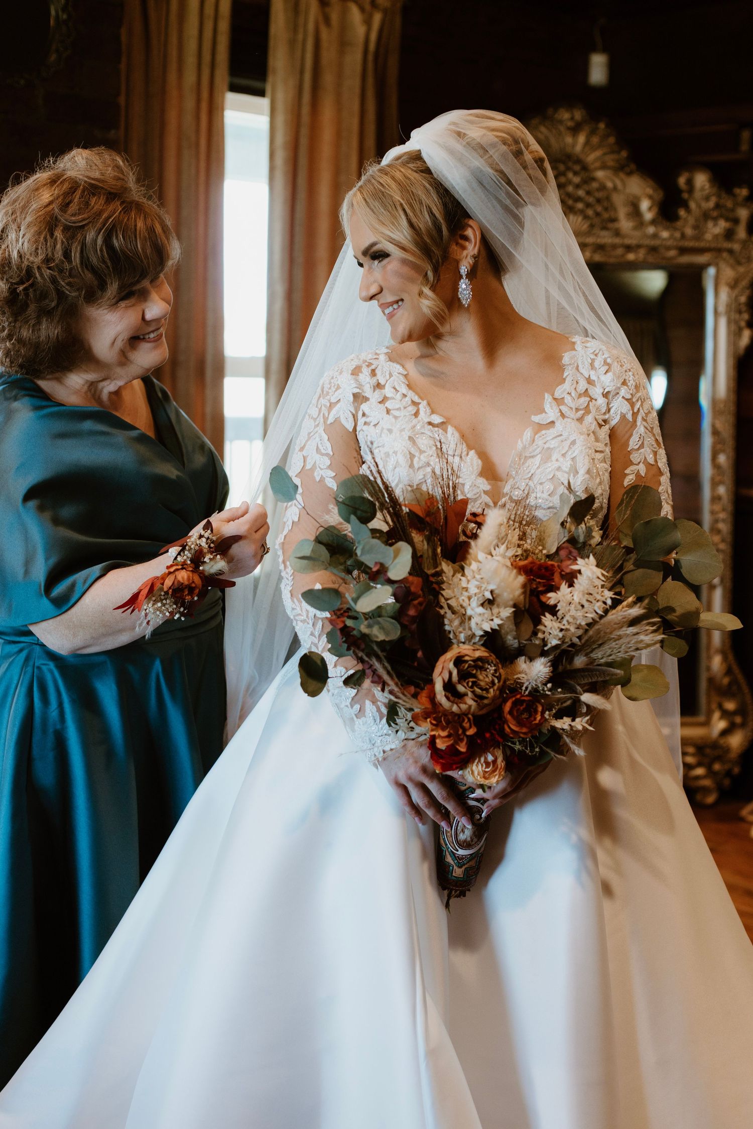 Bride in lace gown holding autumn bouquet smiles as older woman in teal dress helps her prepare for wedding ceremony.
