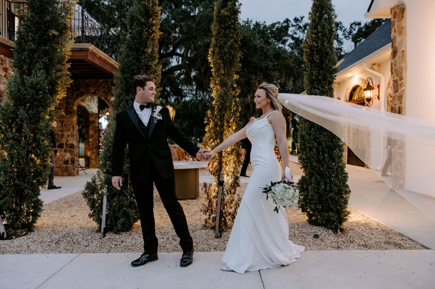 Bride in a white gown with a flowing veil holds hands with a groom in a black tuxedo outdoors beside tall cypress trees at a wedding venue.