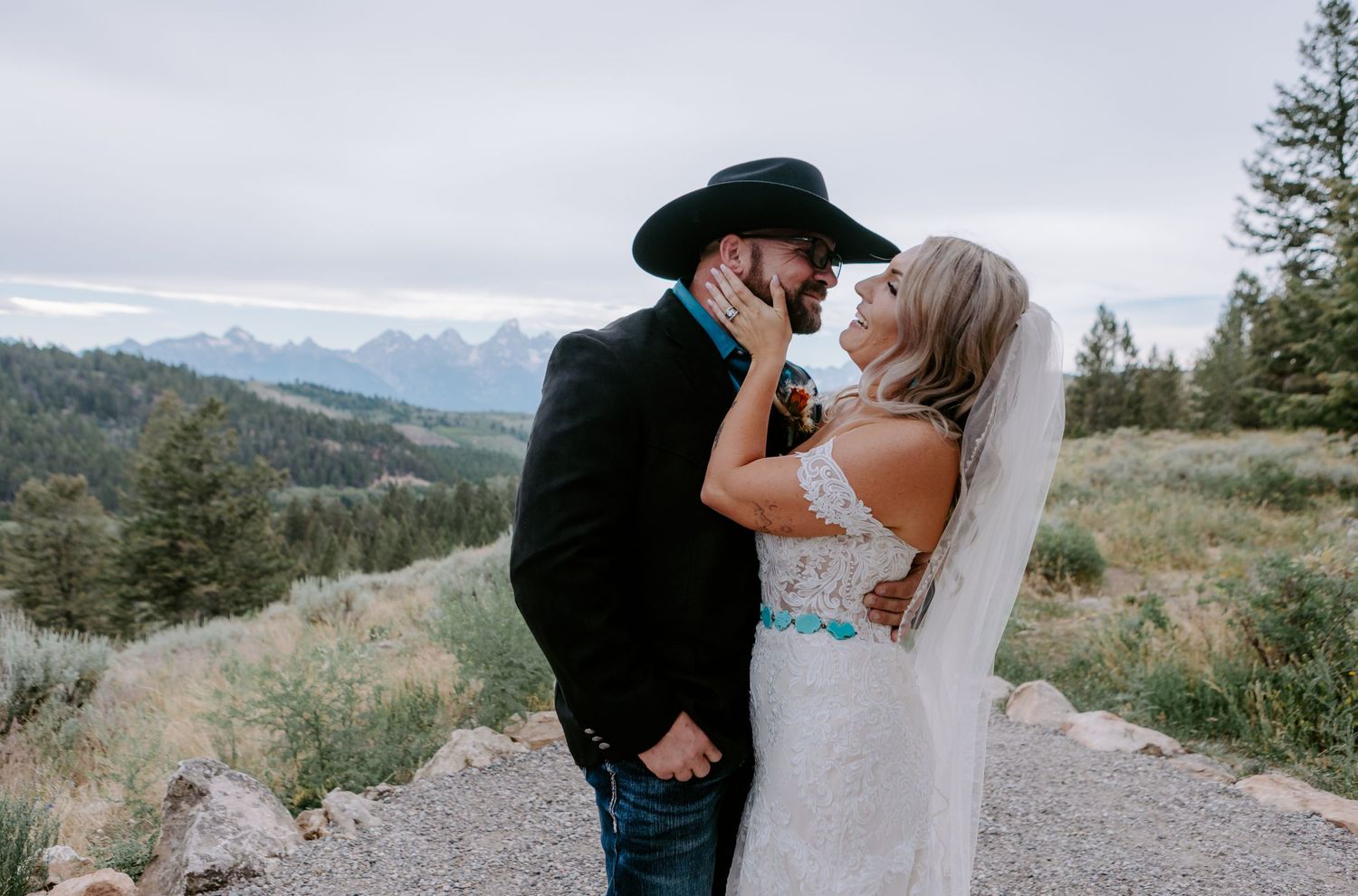 A bride and groom share a romantic moment outdoors with mountain views, the groom wearing a black cowboy hat.