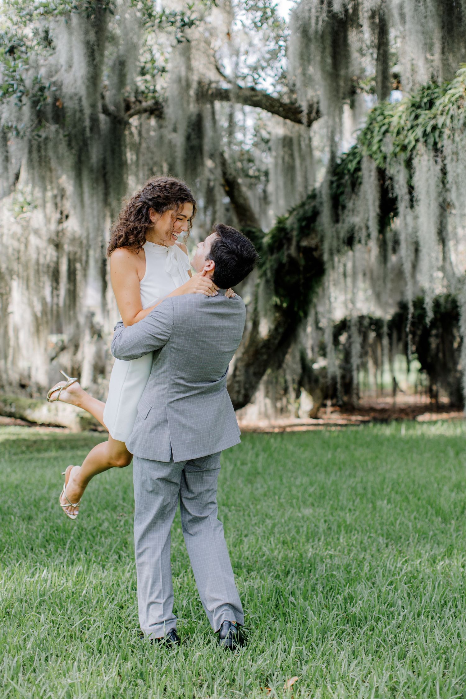 A couple in gray formal attire dances playfully on a lush lawn beneath Spanish moss-draped trees in a Southern garden setting.