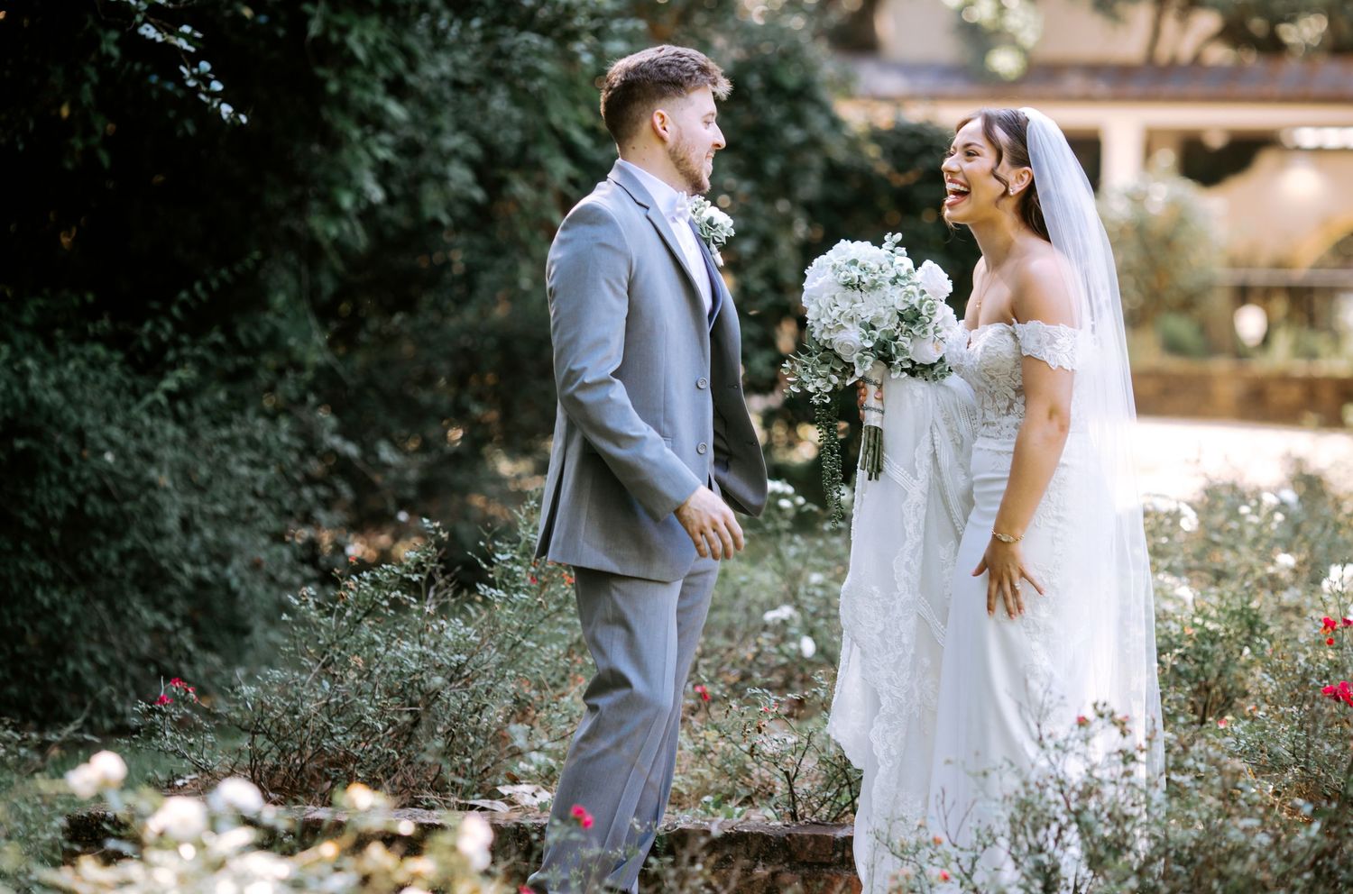 Bride and groom laughing together outdoors in a lush garden, she in a lace off-shoulder gown, he in a gray suit.