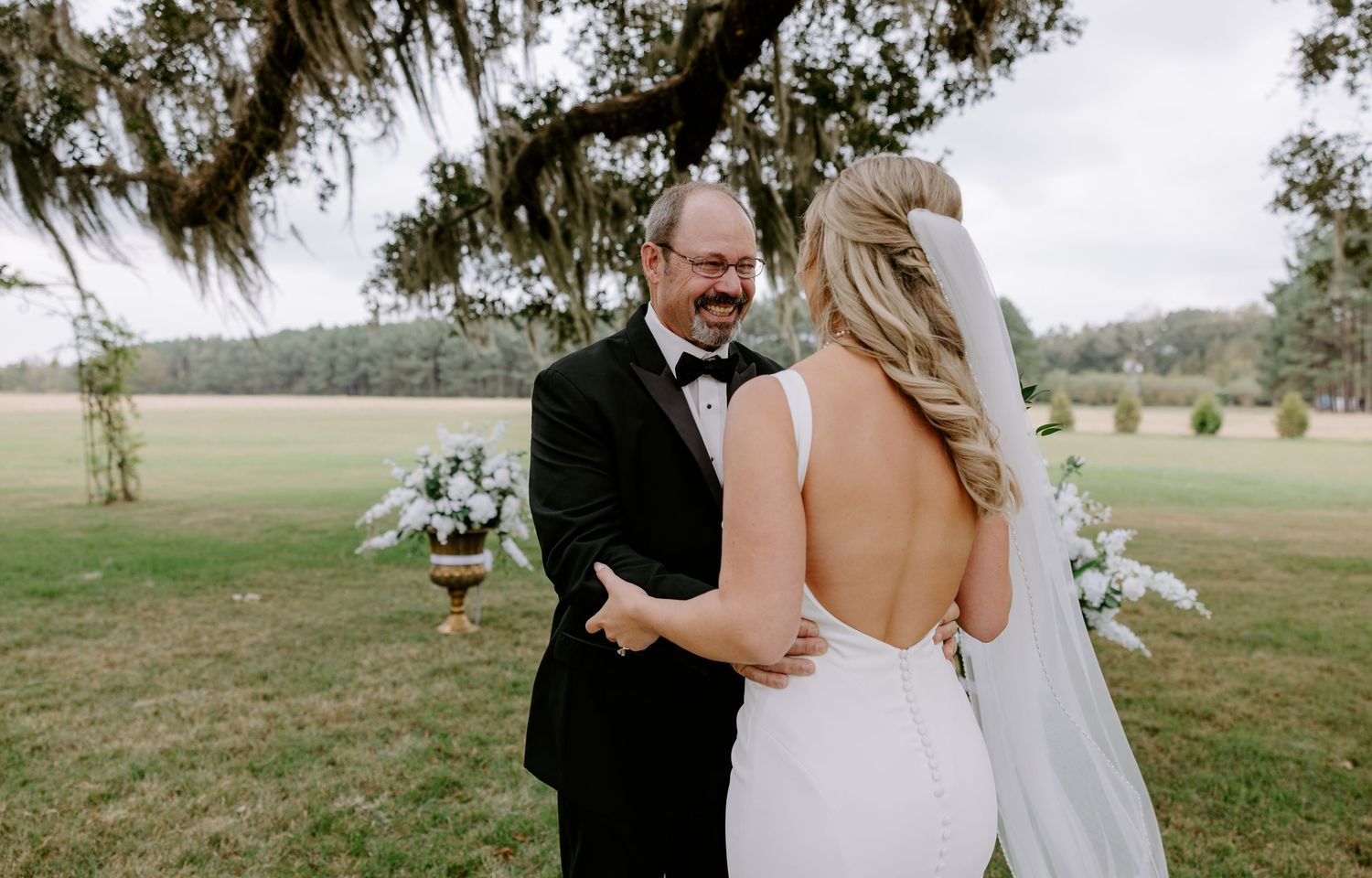 Smiling groom in black tuxedo and bow tie sees his bride in a white gown and veil during a first look under oak trees at an outdoor wedding.