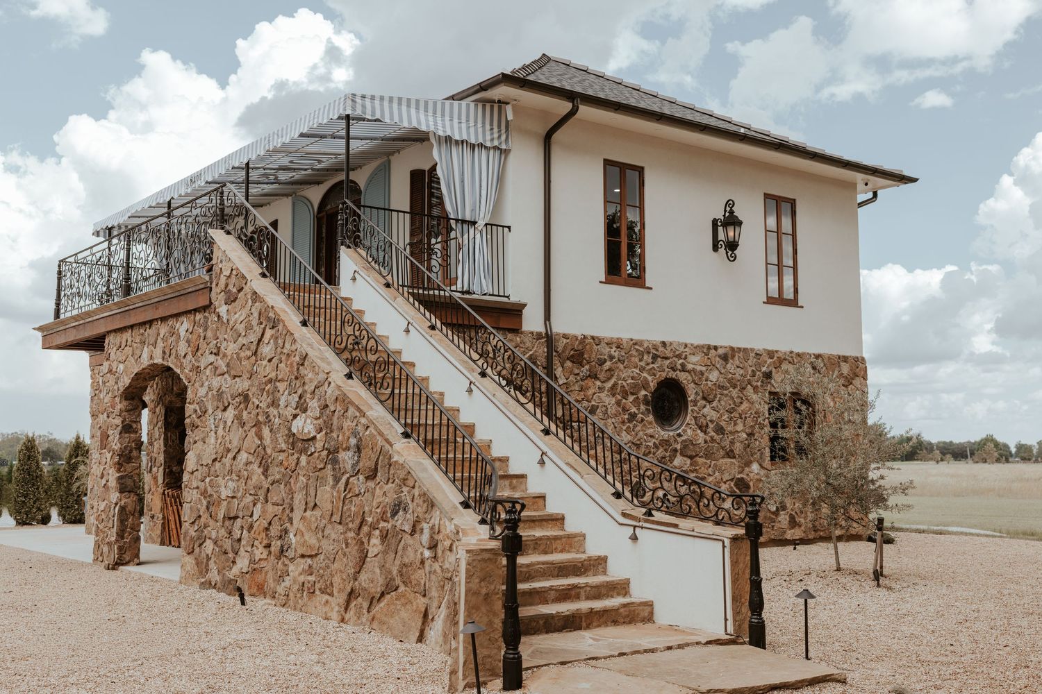Two-story stone and white stucco country house with a wide stone staircase, wrought iron railings, and a covered balcony under a cloudy sky.